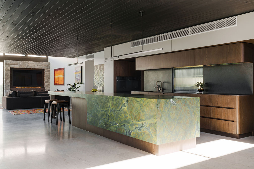 The kitchen at Mezzanine House combines rich timber cabinetry, a dramatic green stone island, dark timber ceilings and an open connection to the living area.