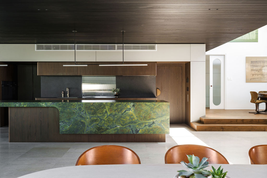 The kitchen at Mezzanine House combines rich timber cabinetry, a dramatic green stone island, dark timber ceilings and an open connection to the living area.