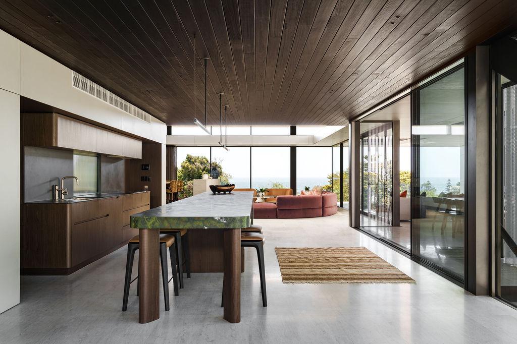 The kitchen at Mezzanine House combines rich timber cabinetry, a dramatic green stone island, dark timber ceilings and an open connection to the living area.