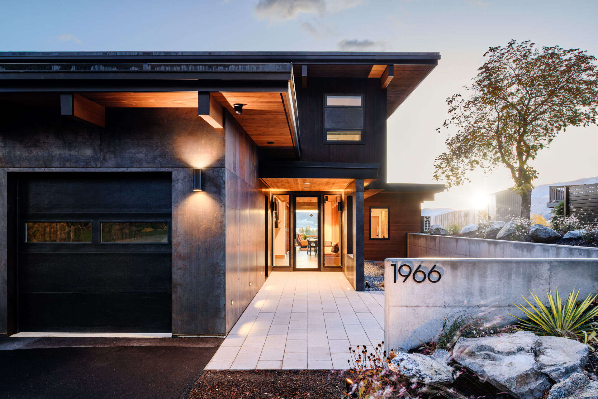 A modern home entrance features a broad paved walkway, glass-panelled front door, deep roof overhangs, natural rock accents, and subtle exterior lighting.