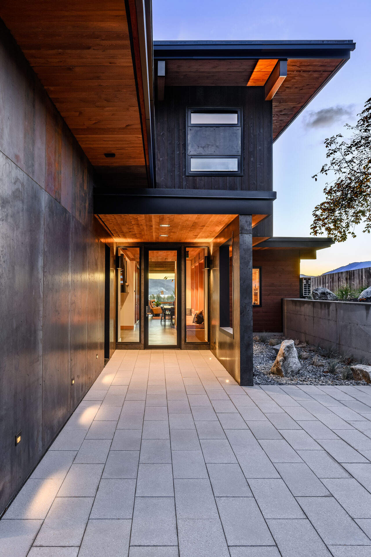 A modern home entrance features a broad paved walkway, glass-panelled front door, deep roof overhangs, natural rock accents, and subtle exterior lighting.