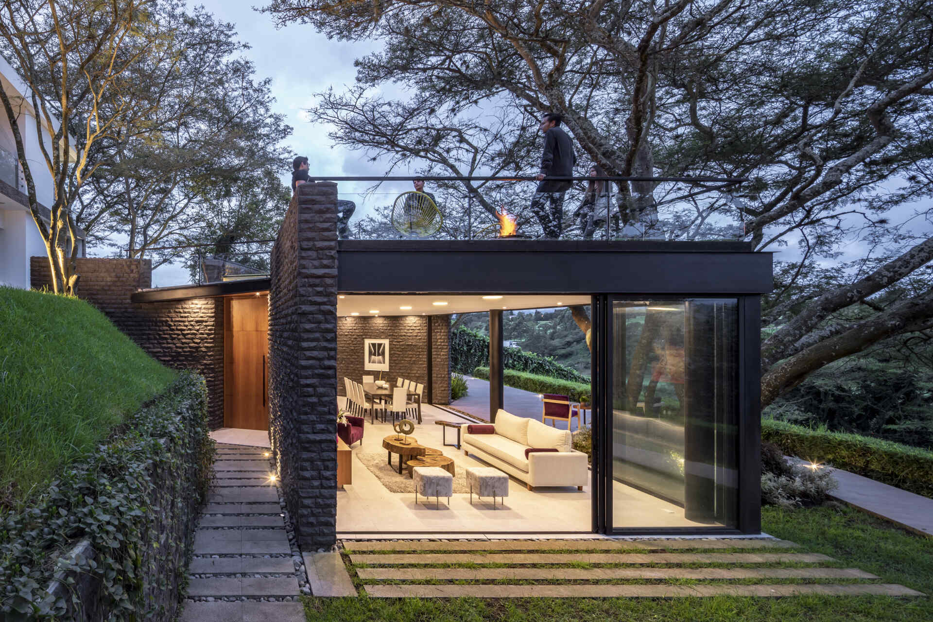 Step inside the Hidden Stone House living room, where floor-to-ceiling glass, dark stone walls and pale furniture look out towards the garden and mature trees.
