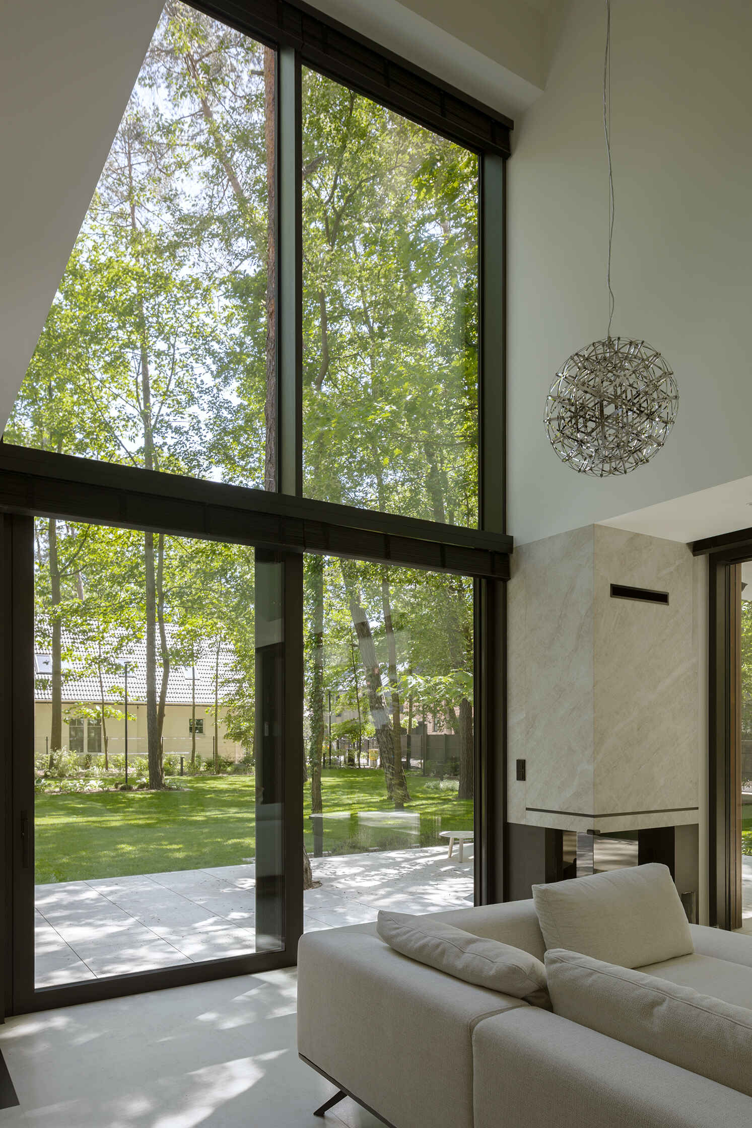 The double-height living room at The Intersecting House connects both levels of the home while opening toward the garden through expansive glazing. The space brings together daylight, forest views and direct terrace access.