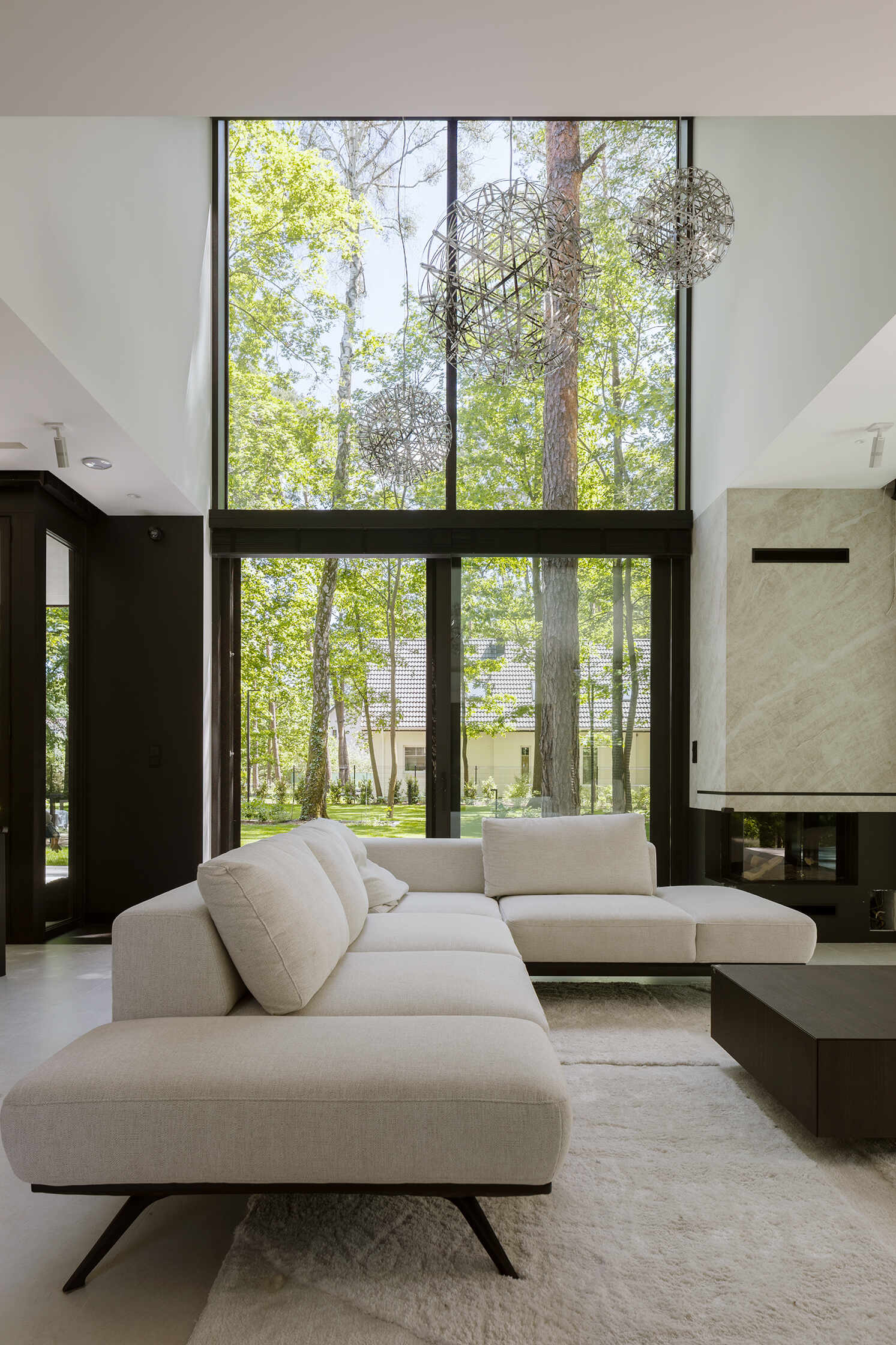 The double-height living room at The Intersecting House connects both levels of the home while opening toward the garden through expansive glazing. The space brings together daylight, forest views and direct terrace access.