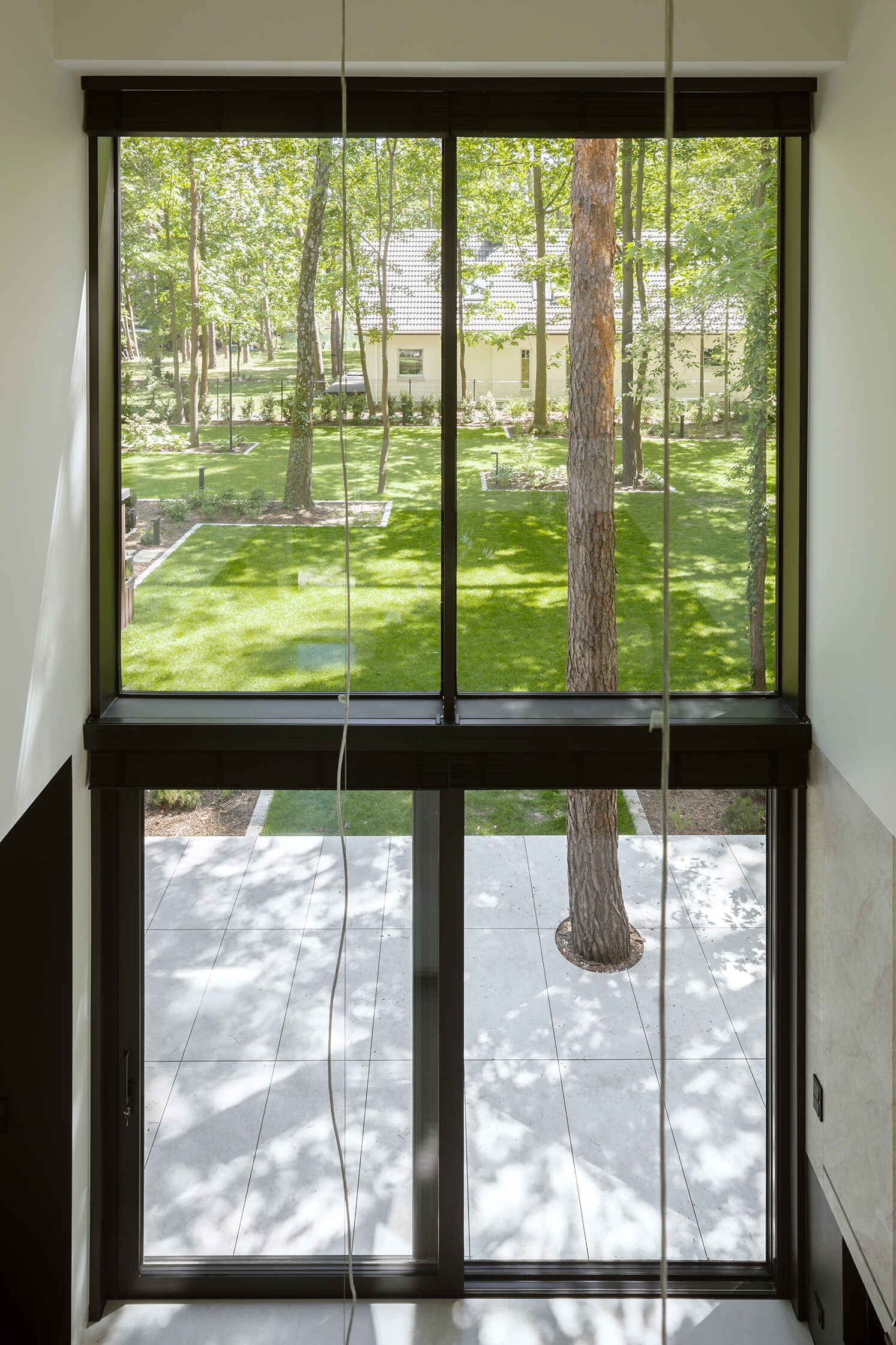 The double-height living room at The Intersecting House connects both levels of the home while opening toward the garden through expansive glazing. The space brings together daylight, forest views and direct terrace access.