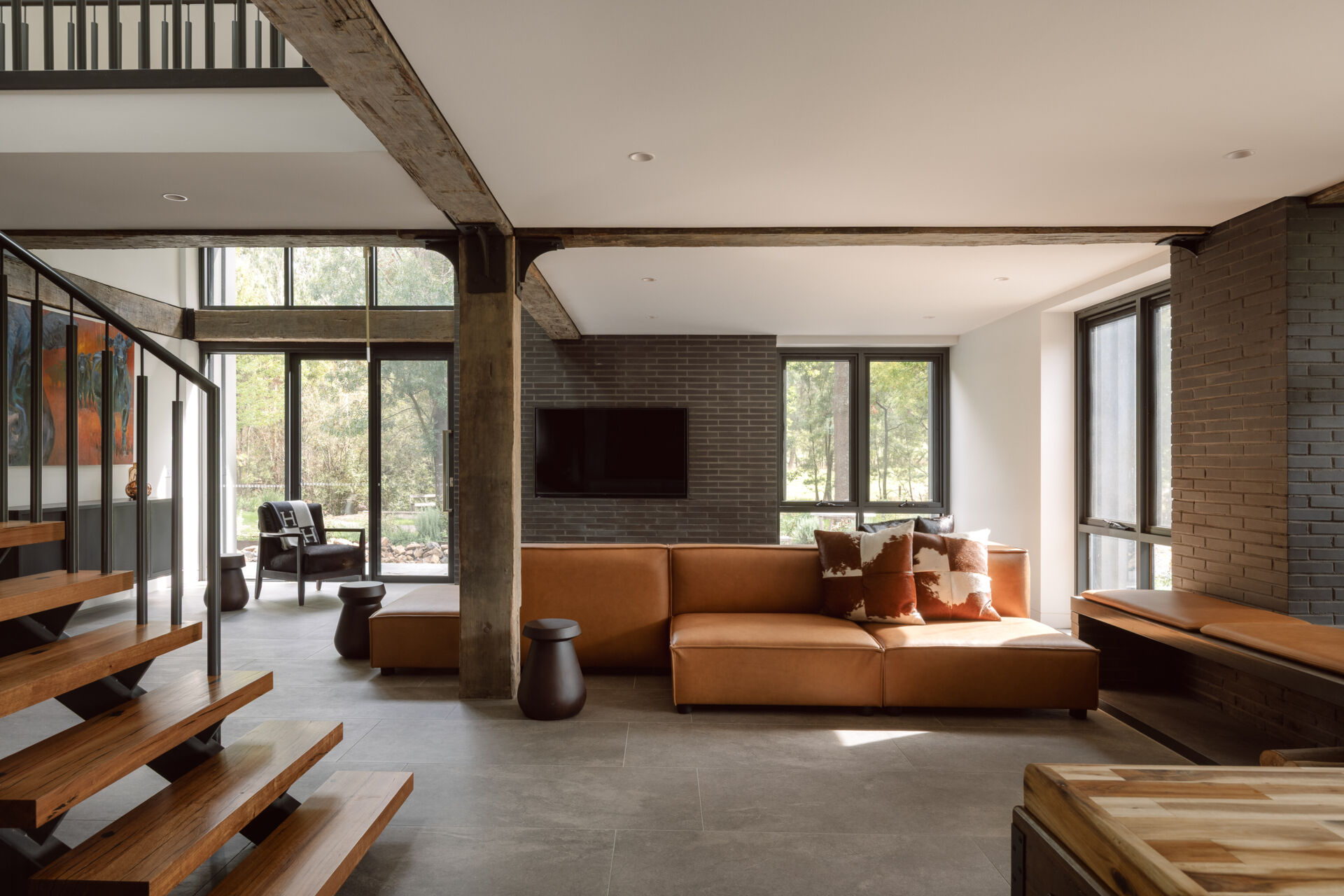 Step inside the living room of Yarra Valley House, featuring a large sectional, dark brick, exposed timber beams, grey flooring and expansive views.
