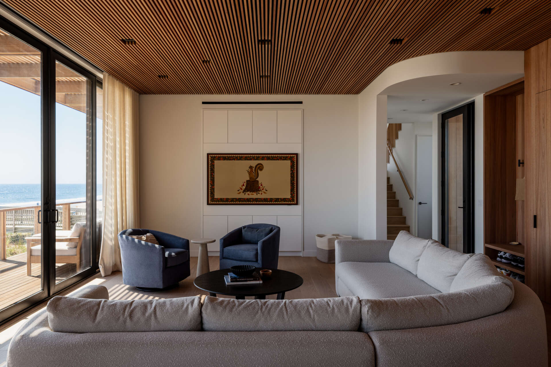 Ocean views, blue armchairs, and pale curved sofas give this Fire Island living room a relaxed coastal feel. Large sliding glass doors open onto a deck, with sheer curtains filtering the daylight.