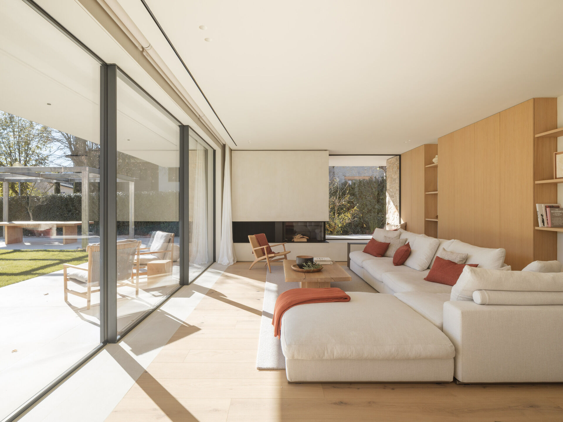 A cream sectional, timber cabinetry, rust-coloured accents, and a fireplace with a window seat furnish this bright living room. Full-height glazing opens the space to garden views at House 2194 by Nordest Architecture SLP in Olot, Spain.