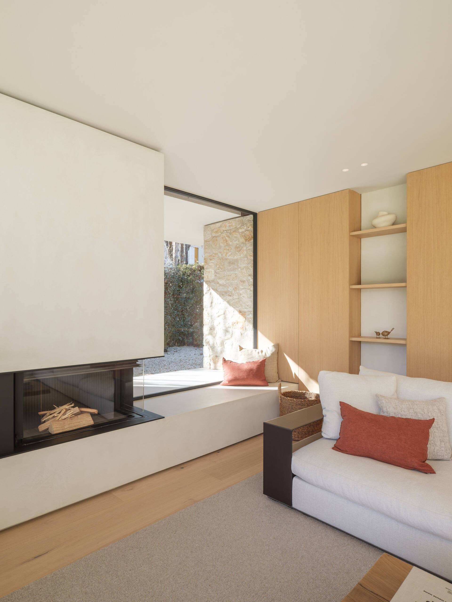 A cream sectional, timber cabinetry, rust-coloured accents, and a fireplace with a window seat furnish this bright living room. Full-height glazing opens the space to garden views at House 2194 by Nordest Architecture SLP in Olot, Spain.