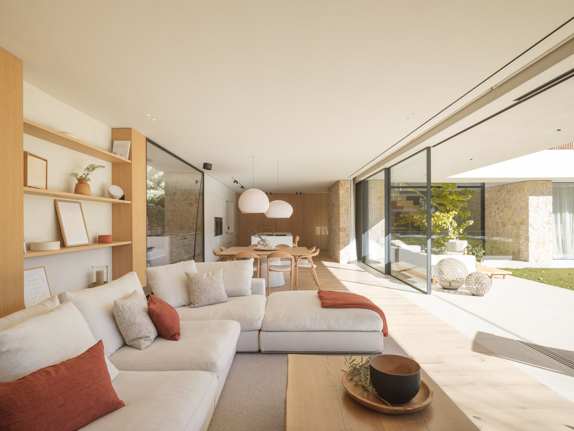 A cream sectional, timber cabinetry, rust-coloured accents, and a fireplace with a window seat furnish this bright living room. Full-height glazing opens the space to garden views at House 2194 by Nordest Architecture SLP in Olot, Spain.