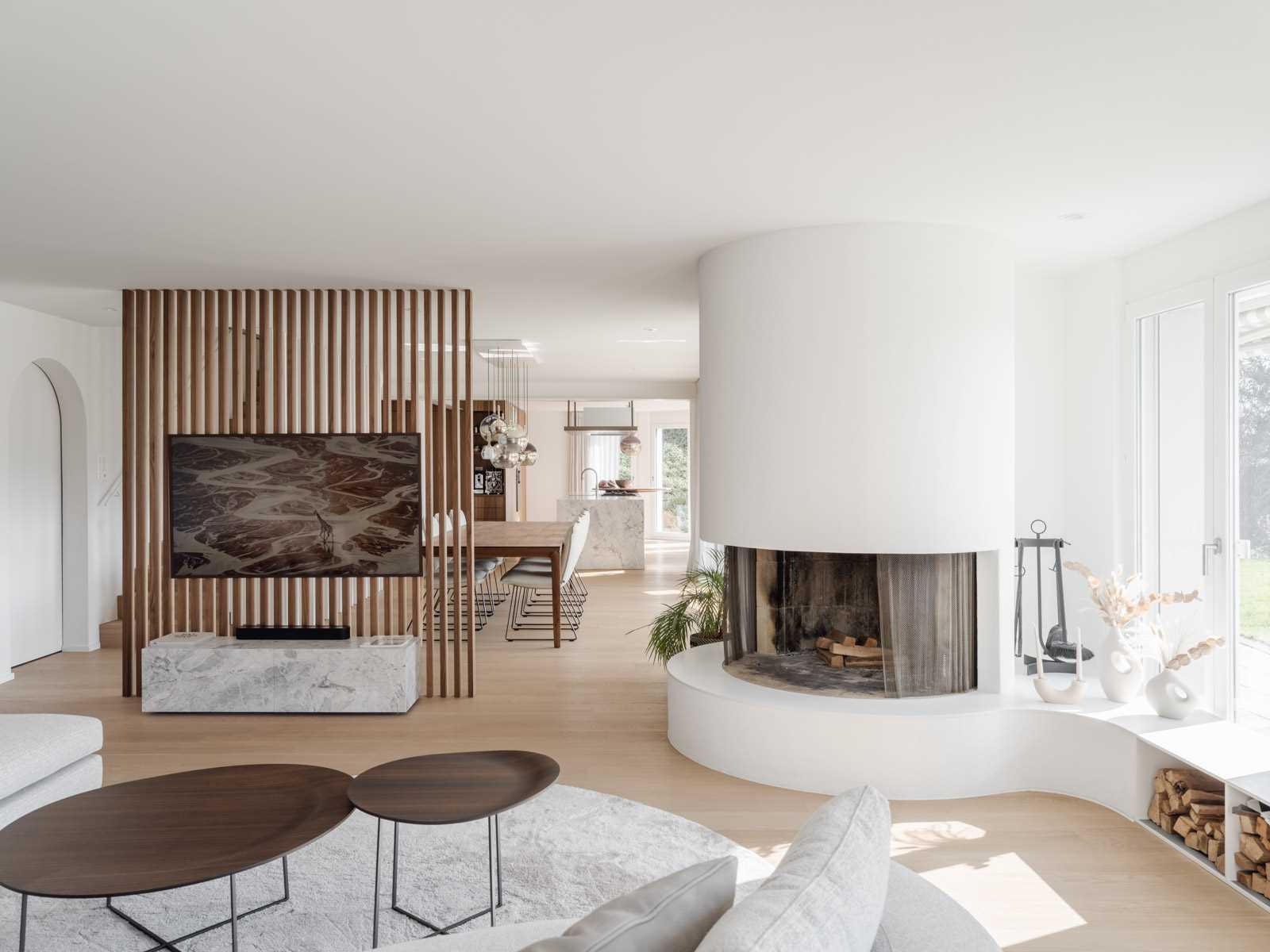 A warm and modern Swiss living room featuring a rounded fireplace, rich hardwood flooring and a wood-slat TV wall that separates the space from the dining area.