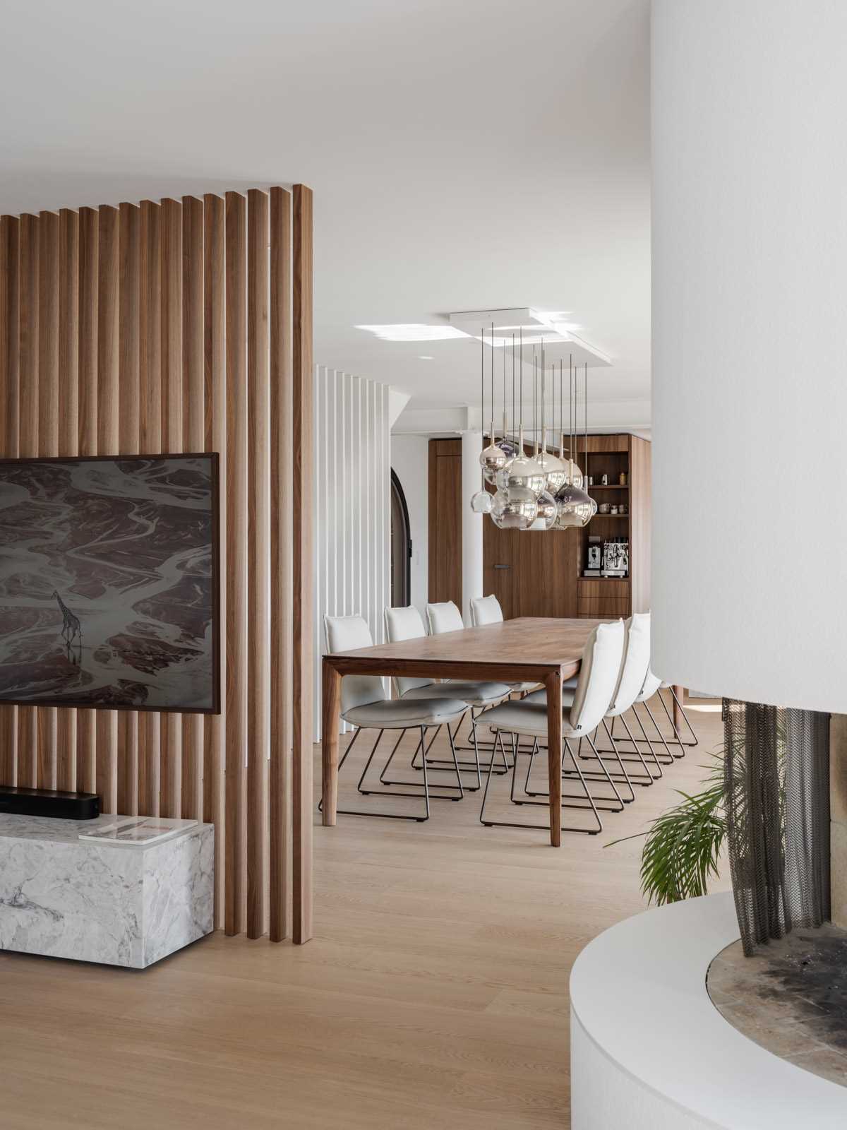 A warm and modern Swiss living room featuring a rounded fireplace, rich hardwood flooring and a wood-slat TV wall that separates the space from the dining area.