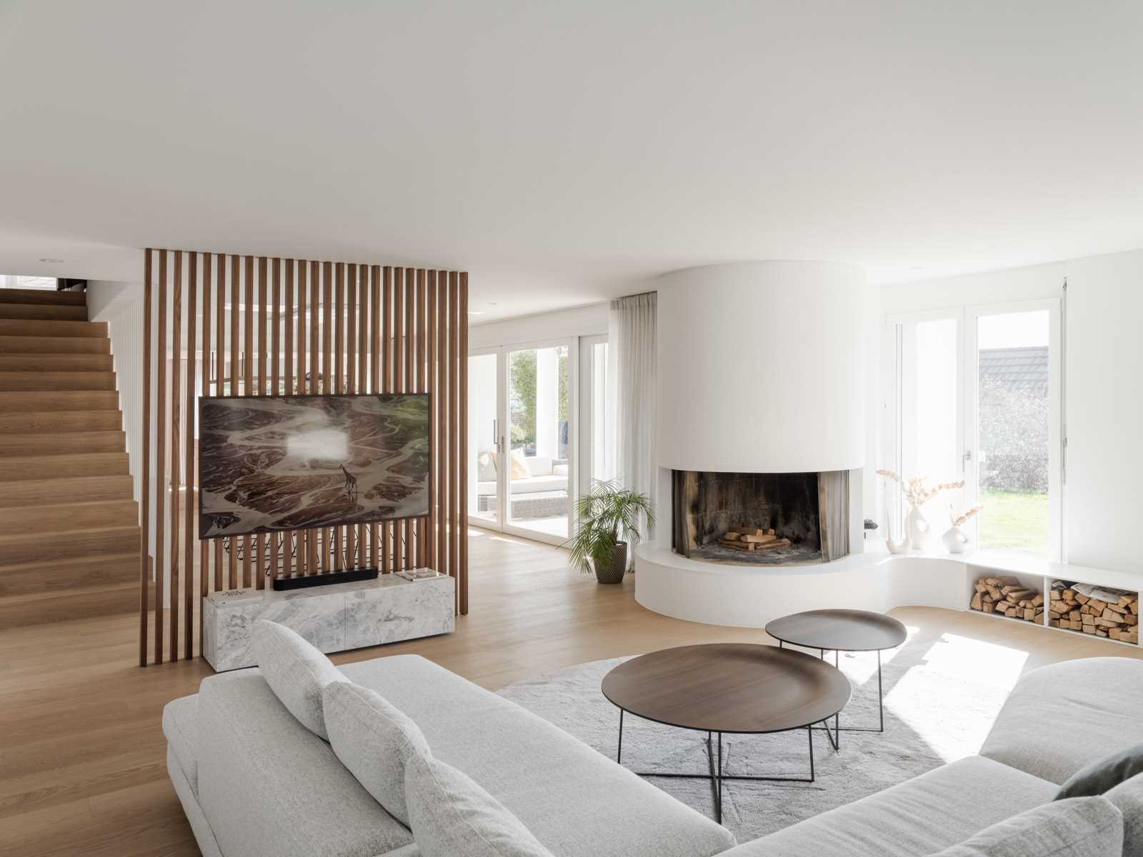 A warm and modern Swiss living room featuring a rounded fireplace, rich hardwood flooring and a wood-slat TV wall that separates the space from the dining area.