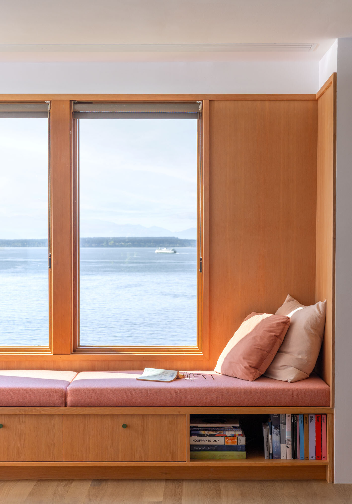 Step inside the renovated living room at Alki Lookout, where pale timber, pink upholstery, patterned tile and a long built-in window bench create a warm place to sit and watch ferries cross Puget Sound.