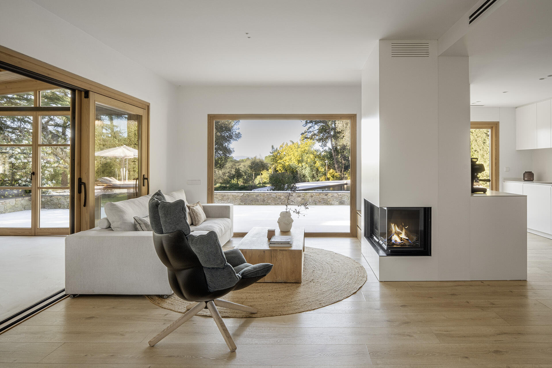 A light-filled living room by Susanna Cots pairs a white Flexform sofa, sculptural seating and pale timber flooring with large framed views of the forest. A glass-fronted fireplace separates the kitchen and lounge without closing off the space.