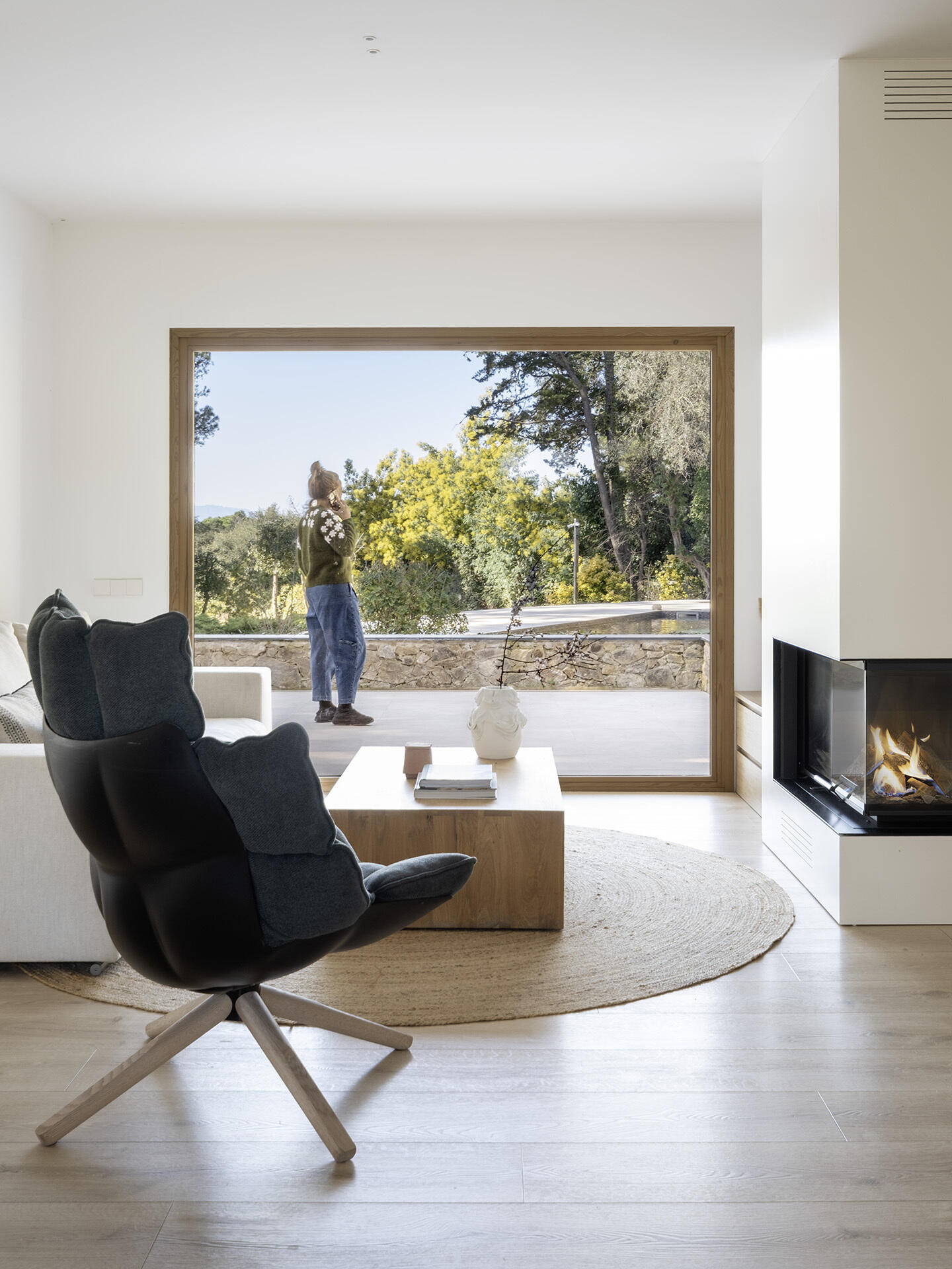 A light-filled living room by Susanna Cots pairs a white Flexform sofa, sculptural seating and pale timber flooring with large framed views of the forest. A glass-fronted fireplace separates the kitchen and lounge without closing off the space.