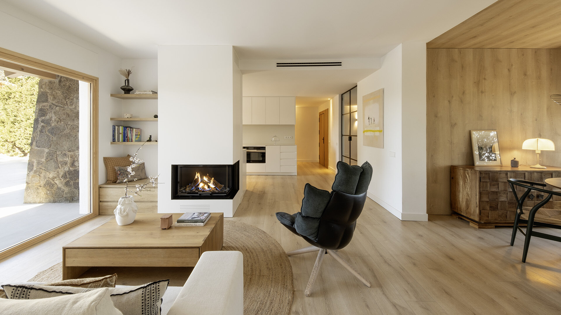 A light-filled living room by Susanna Cots pairs a white Flexform sofa, sculptural seating and pale timber flooring with large framed views of the forest. A glass-fronted fireplace separates the kitchen and lounge without closing off the space.