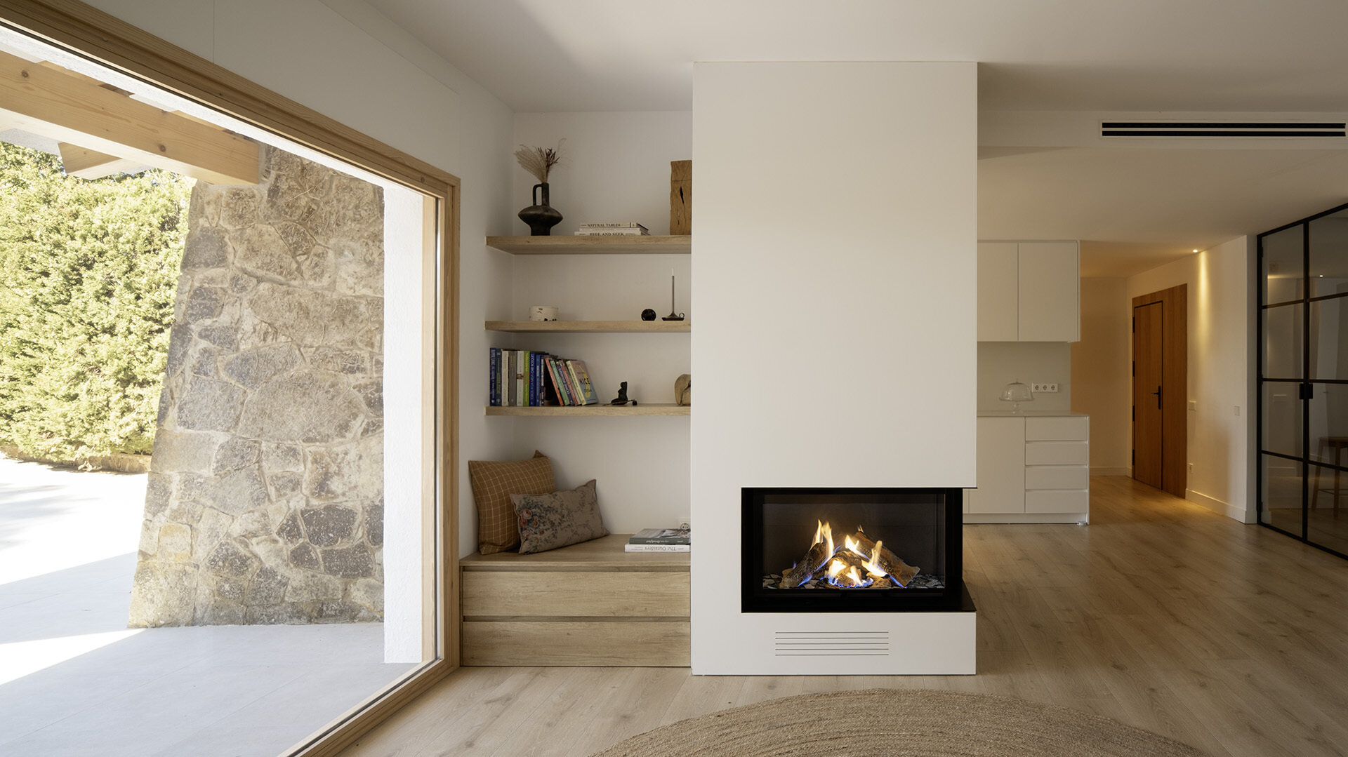A light-filled living room by Susanna Cots pairs a white Flexform sofa, sculptural seating and pale timber flooring with large framed views of the forest. A glass-fronted fireplace separates the kitchen and lounge without closing off the space.