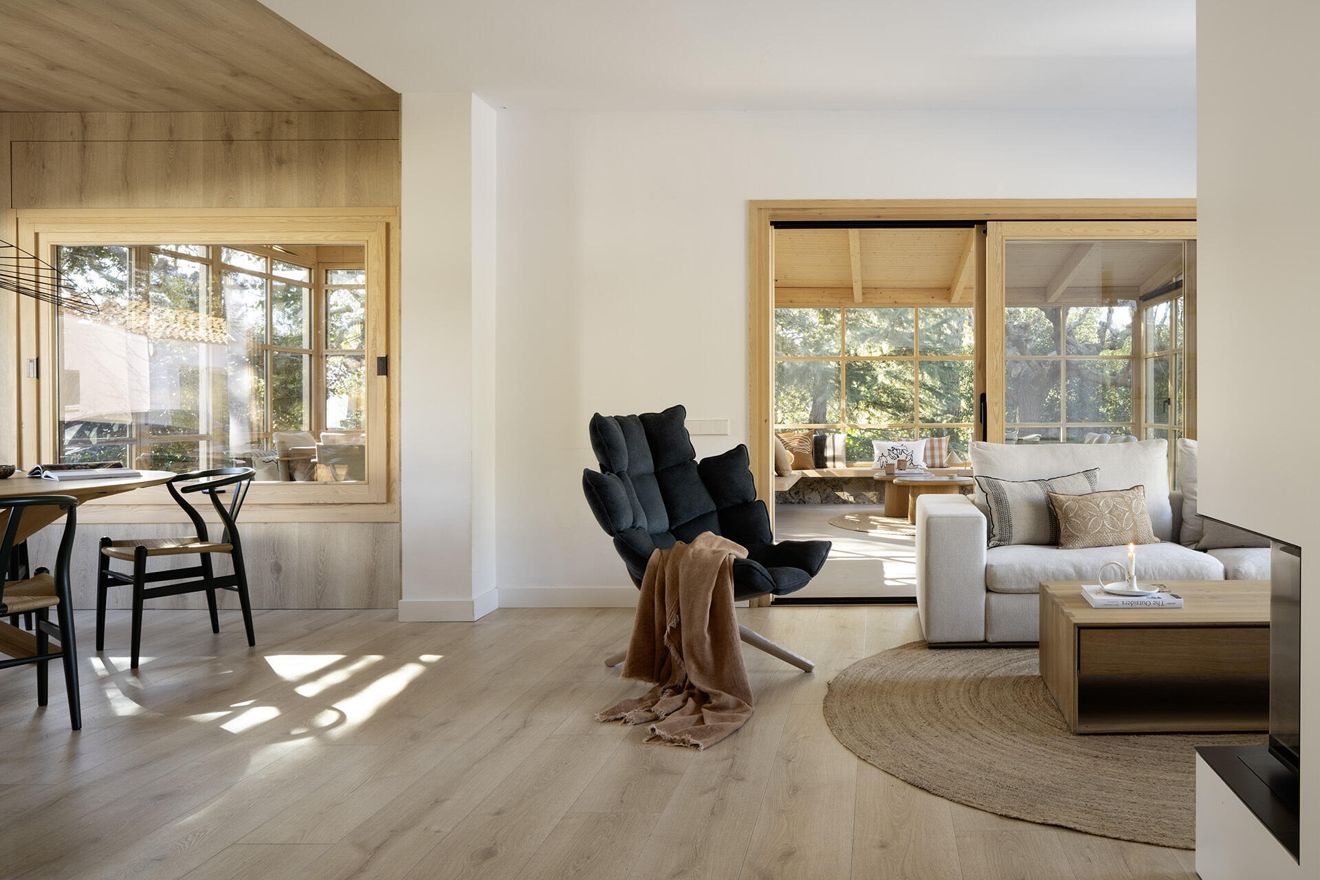 A light-filled living room by Susanna Cots pairs a white Flexform sofa, sculptural seating and pale timber flooring with large framed views of the forest. A glass-fronted fireplace separates the kitchen and lounge without closing off the space.