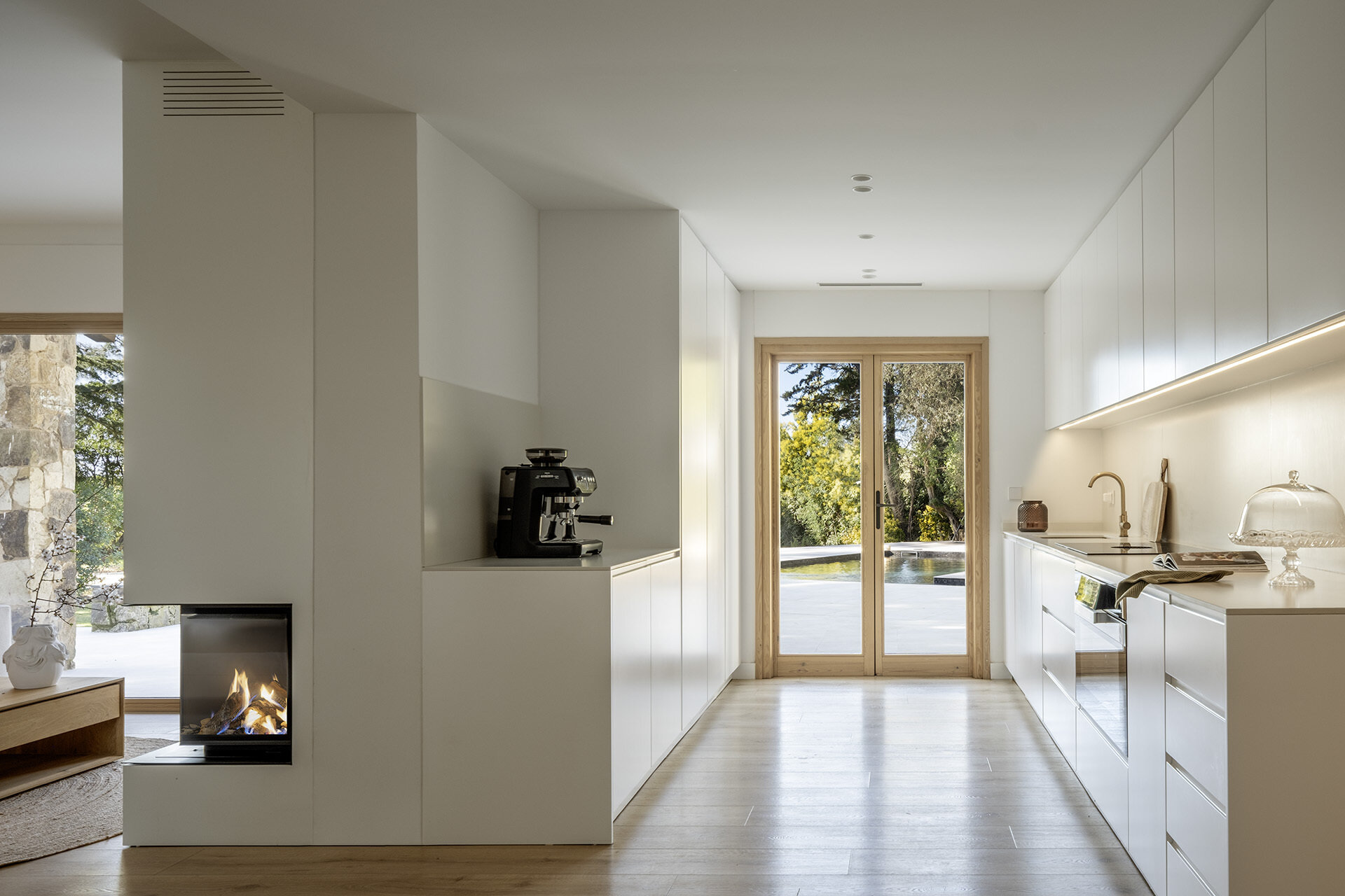 This minimalist white kitchen by Susanna Cots features handle-free cabinetry, pale timber flooring and a direct view toward the pool. Behind black-framed glass doors, a compact home office continues the clean palette with built-in desks and timber-framed windows.