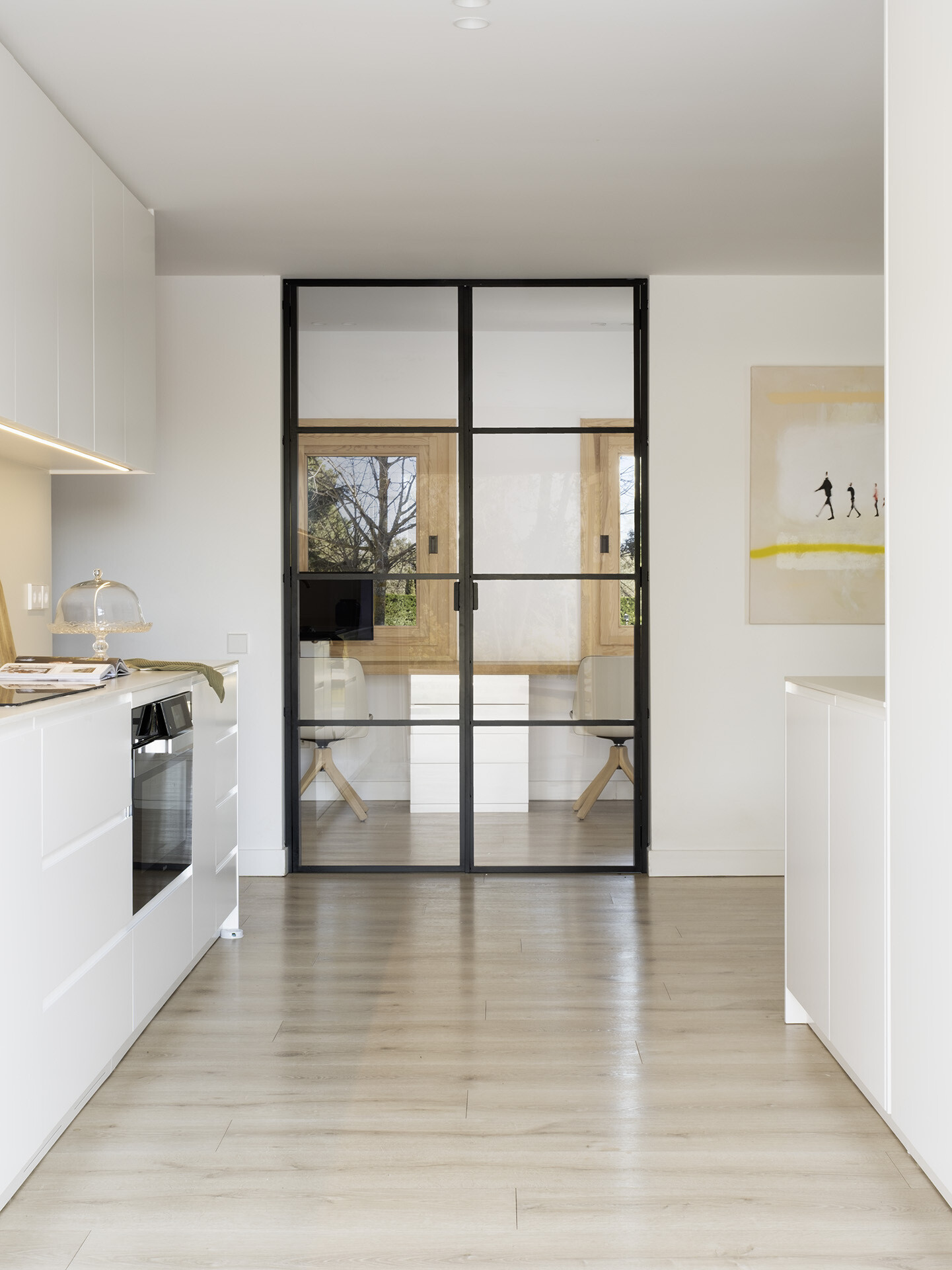 This minimalist white kitchen by Susanna Cots features handle-free cabinetry, pale timber flooring and a direct view toward the pool. Behind black-framed glass doors, a compact home office continues the clean palette with built-in desks and timber-framed windows.