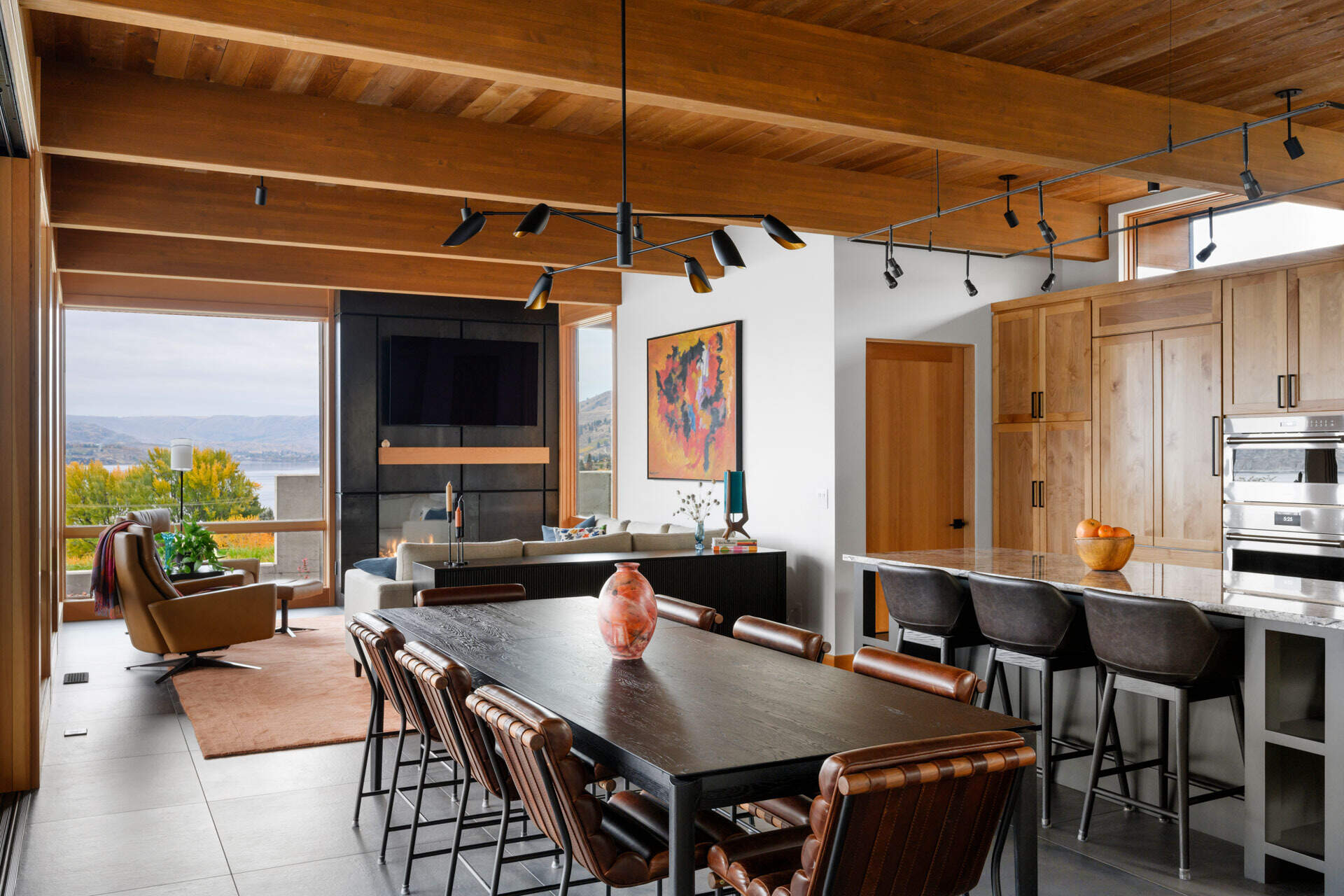 A warm dining area with exposed timber beams, leather chairs, a dark dining table, and expansive lake views, combining natural materials with modern architecture.