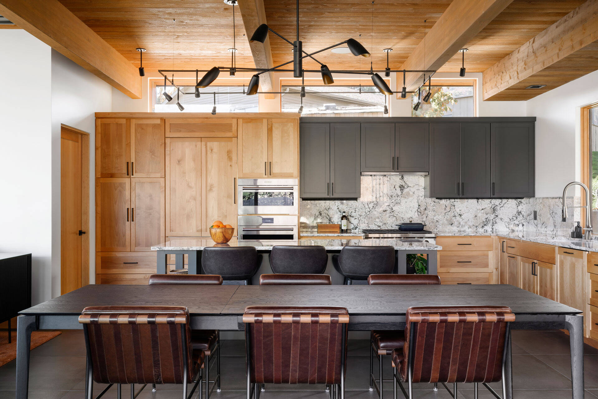 A warm dining area with exposed timber beams, leather chairs, a dark dining table, and expansive lake views, combining natural materials with modern architecture.