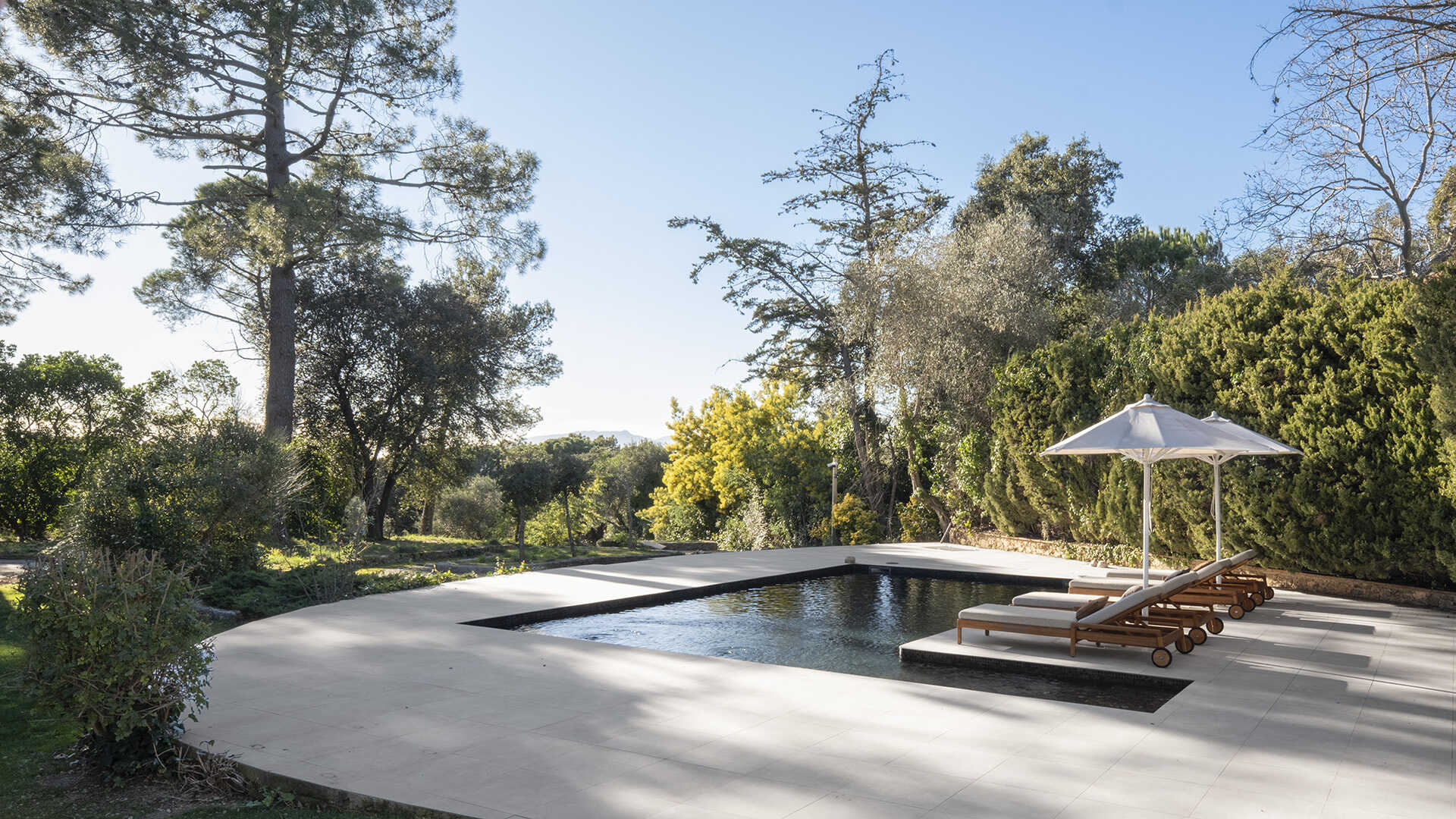 The outdoor spaces include a dark contemporary kitchen, a broad pool terrace and a barrel-shaped timber sauna tucked between mature trees. Together, they create several places for cooking, swimming and relaxing throughout the garden.