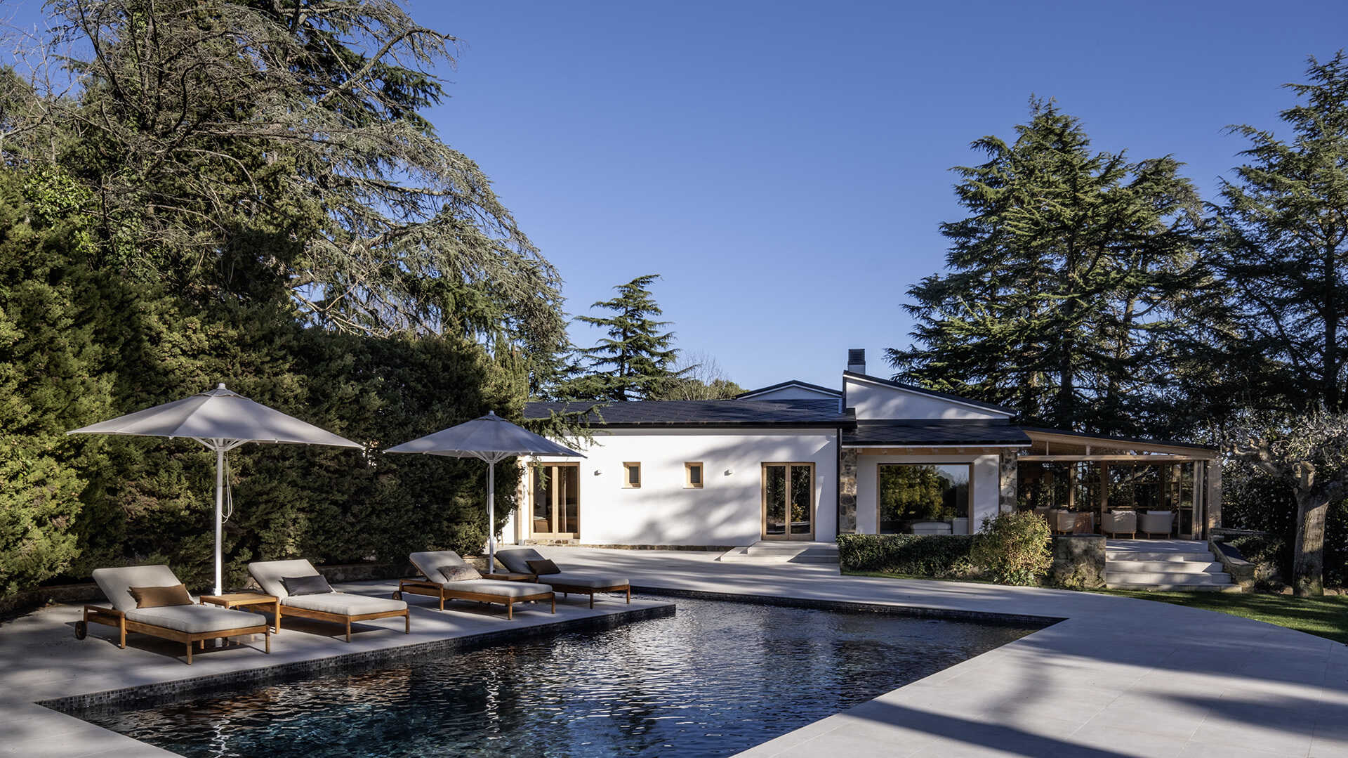 The outdoor spaces include a dark contemporary kitchen, a broad pool terrace and a barrel-shaped timber sauna tucked between mature trees. Together, they create several places for cooking, swimming and relaxing throughout the garden.