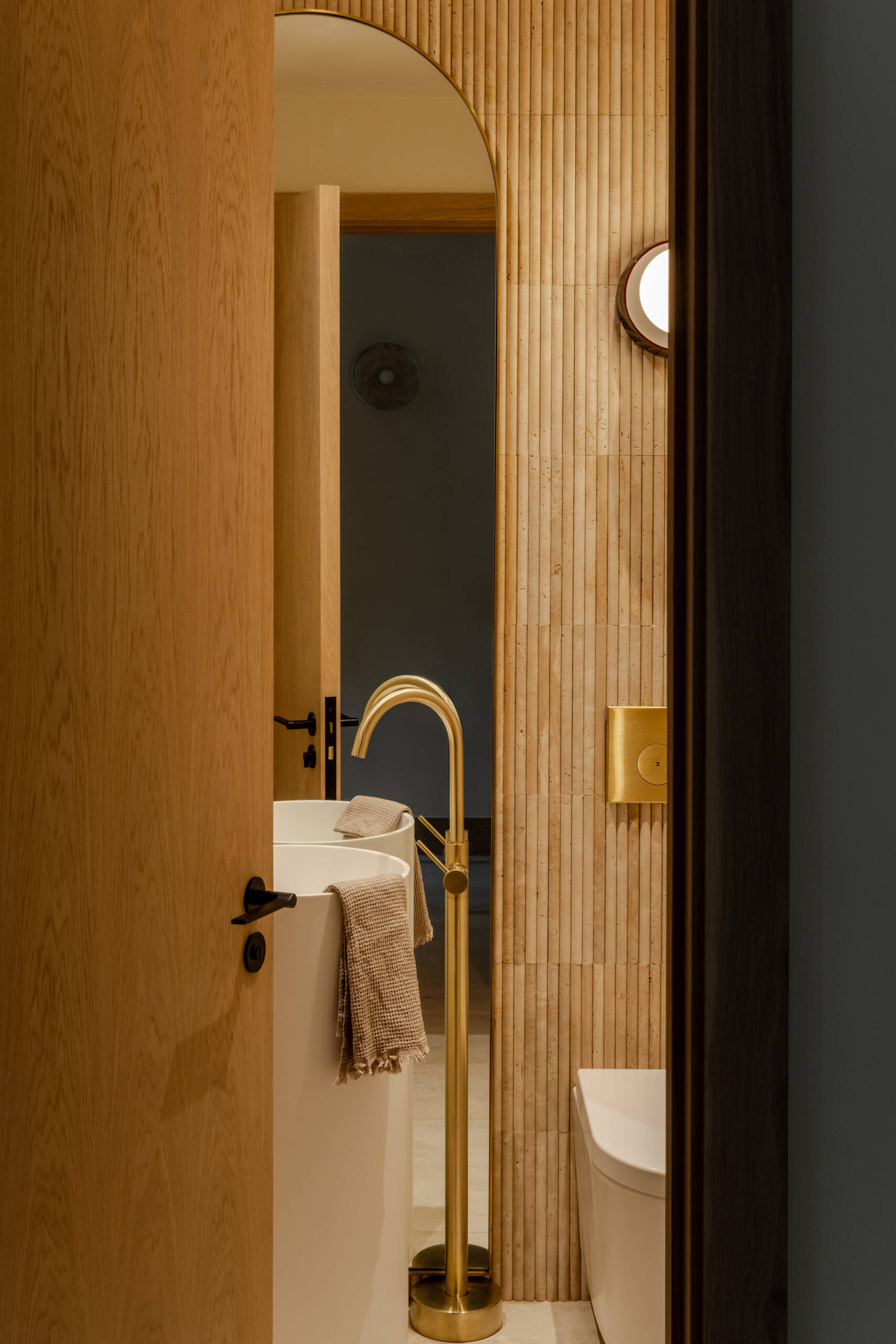 See the compact powder room at Thirsk Cottage, combining fluted stone, warm wood, brushed brass, a freestanding sink and an elegant arched mirror.