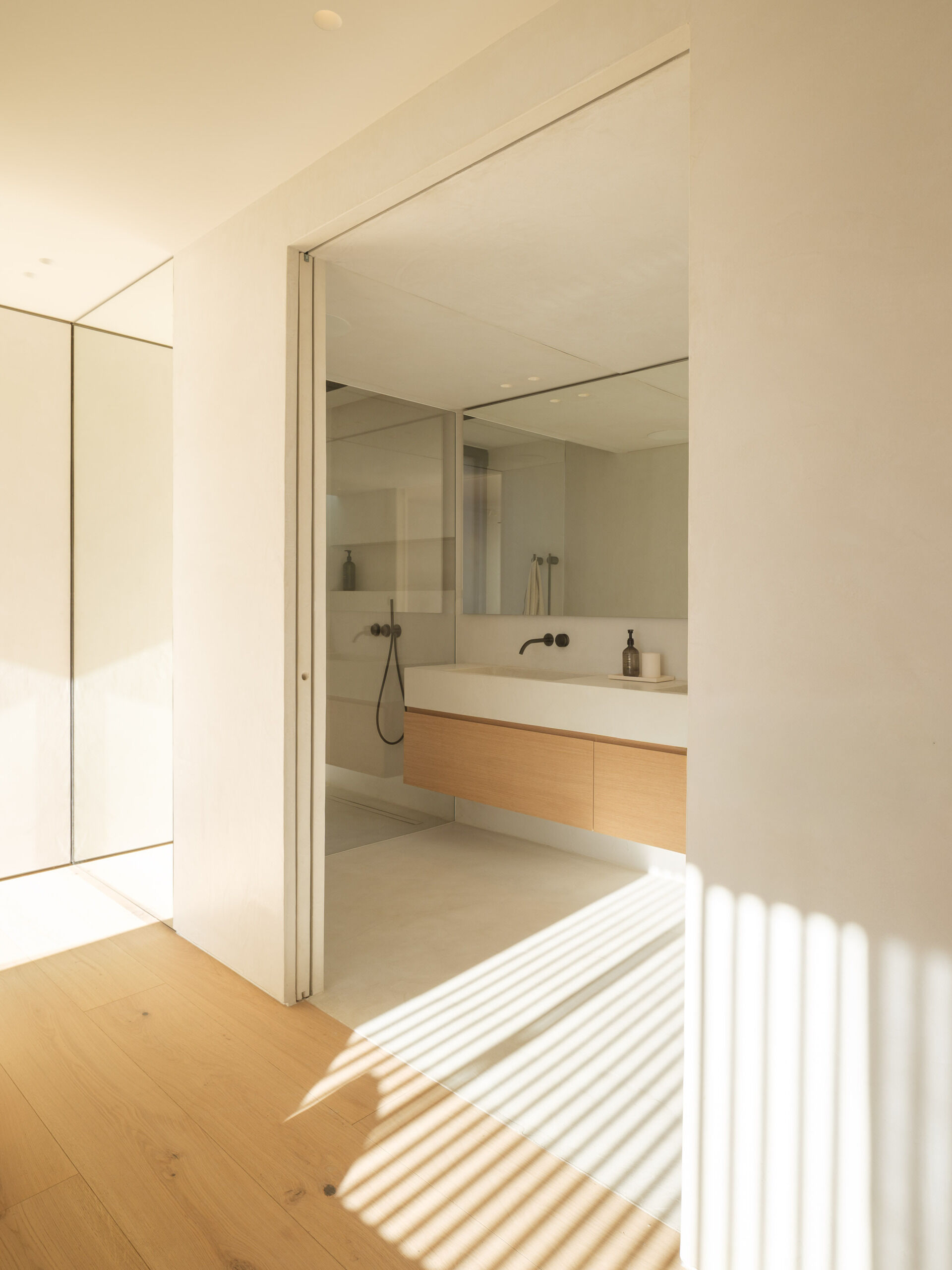 In this bathroom, a broad mirror sits above a substantial rectangular basin, paired with dark wall-mounted taps. A clear glass shower enclosure keeps the compact layout visually open, and a recessed shelf provides space for toiletries. At the doorway, sunlight filtered through the exterior slats draws narrow stripes across the timber floor.