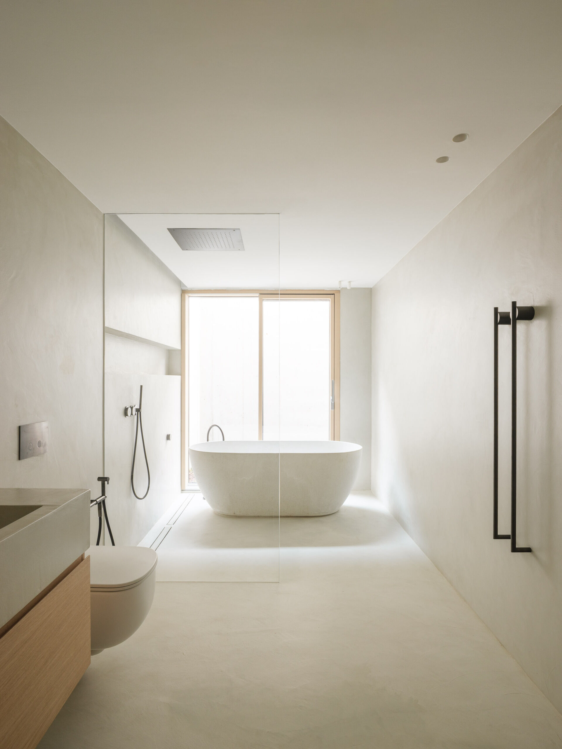 In this bathroom, a freestanding oval bathtub sits in front of a large window at the far end of the room. A frameless glass panel separates the shower area, where a square rainfall head is set into the ceiling and a handheld shower sits beside a long wall niche. Dark fittings and two slender towel rails punctuate the light surfaces, while a wall-hung toilet leaves the floor clear beneath it.