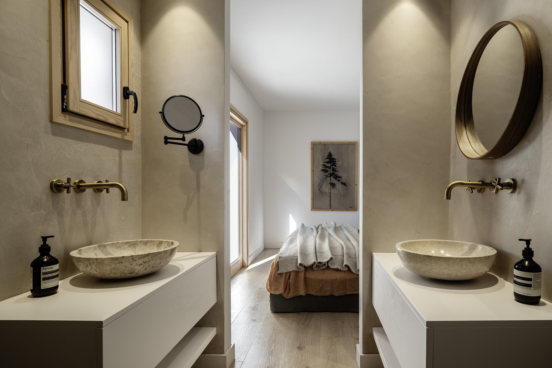 The primary bedroom pairs a Gervasoni bed with warm timber cladding, neutral textiles and direct views toward the pool. The ensuite features twin vanity areas with stone basins, brass taps and timber-framed windows.