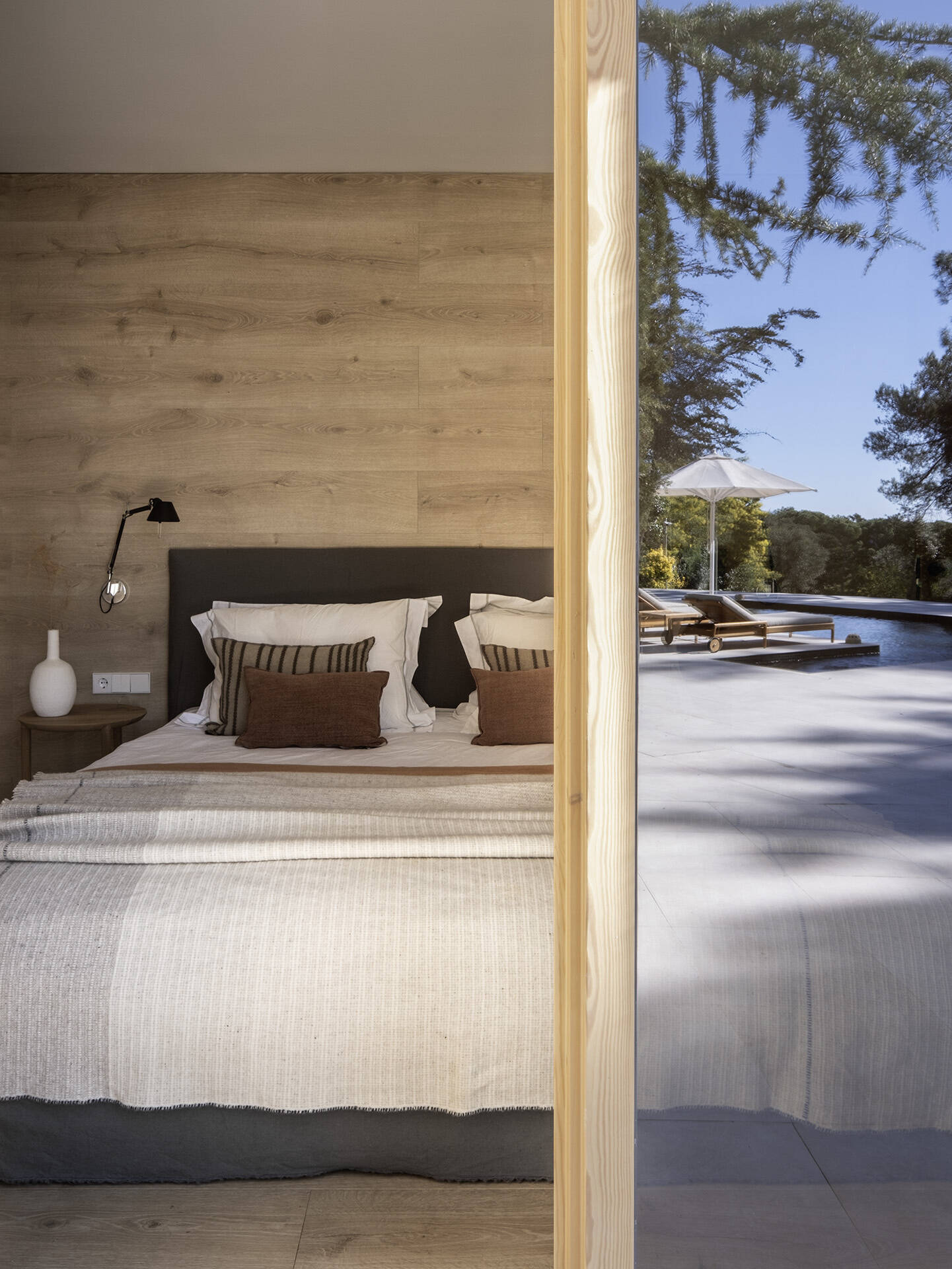 The primary bedroom pairs a Gervasoni bed with warm timber cladding, neutral textiles and direct views toward the pool. The ensuite features twin vanity areas with stone basins, brass taps and timber-framed windows.