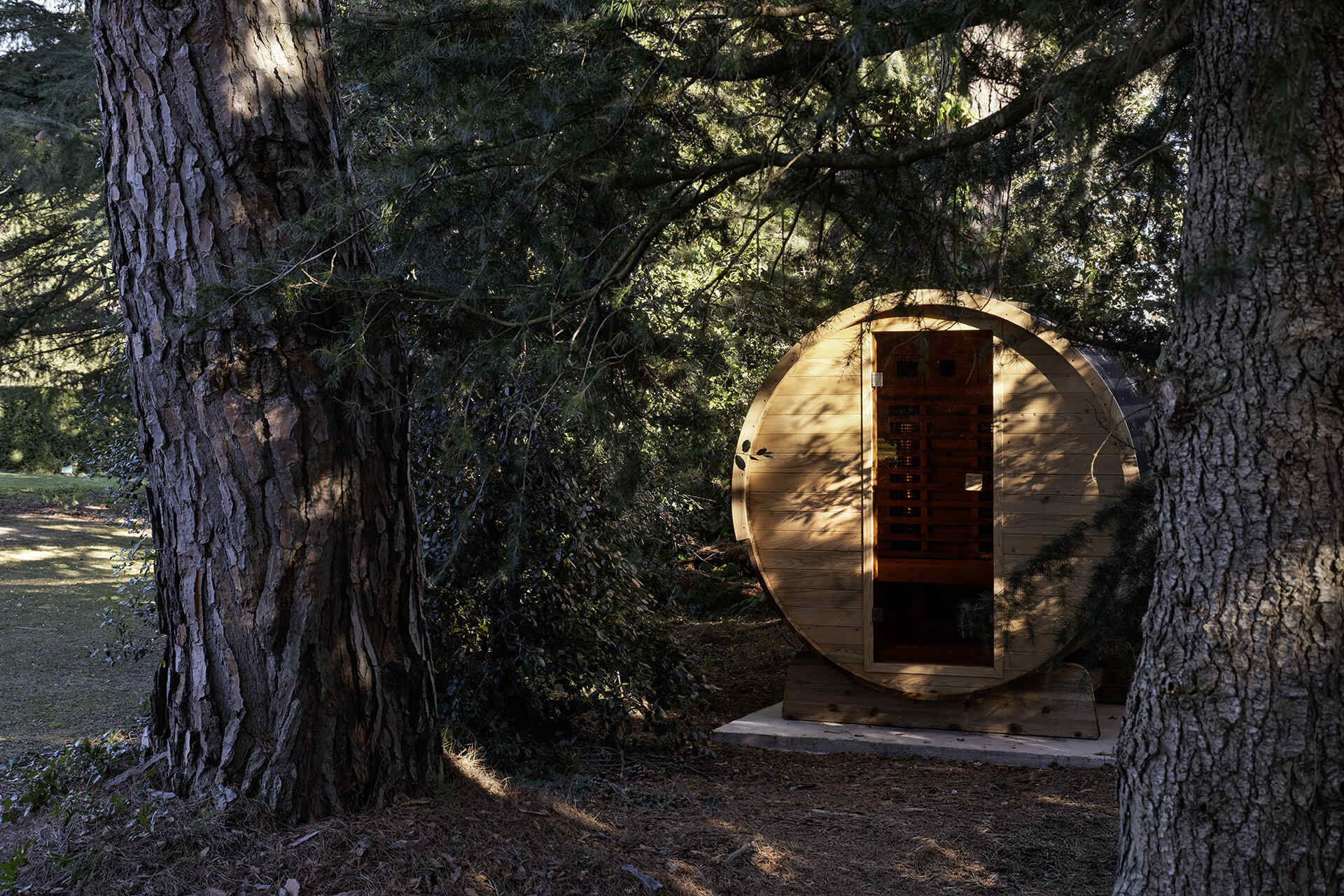 The outdoor spaces include a dark contemporary kitchen, a broad pool terrace and a barrel-shaped timber sauna tucked between mature trees. Together, they create several places for cooking, swimming and relaxing throughout the garden.