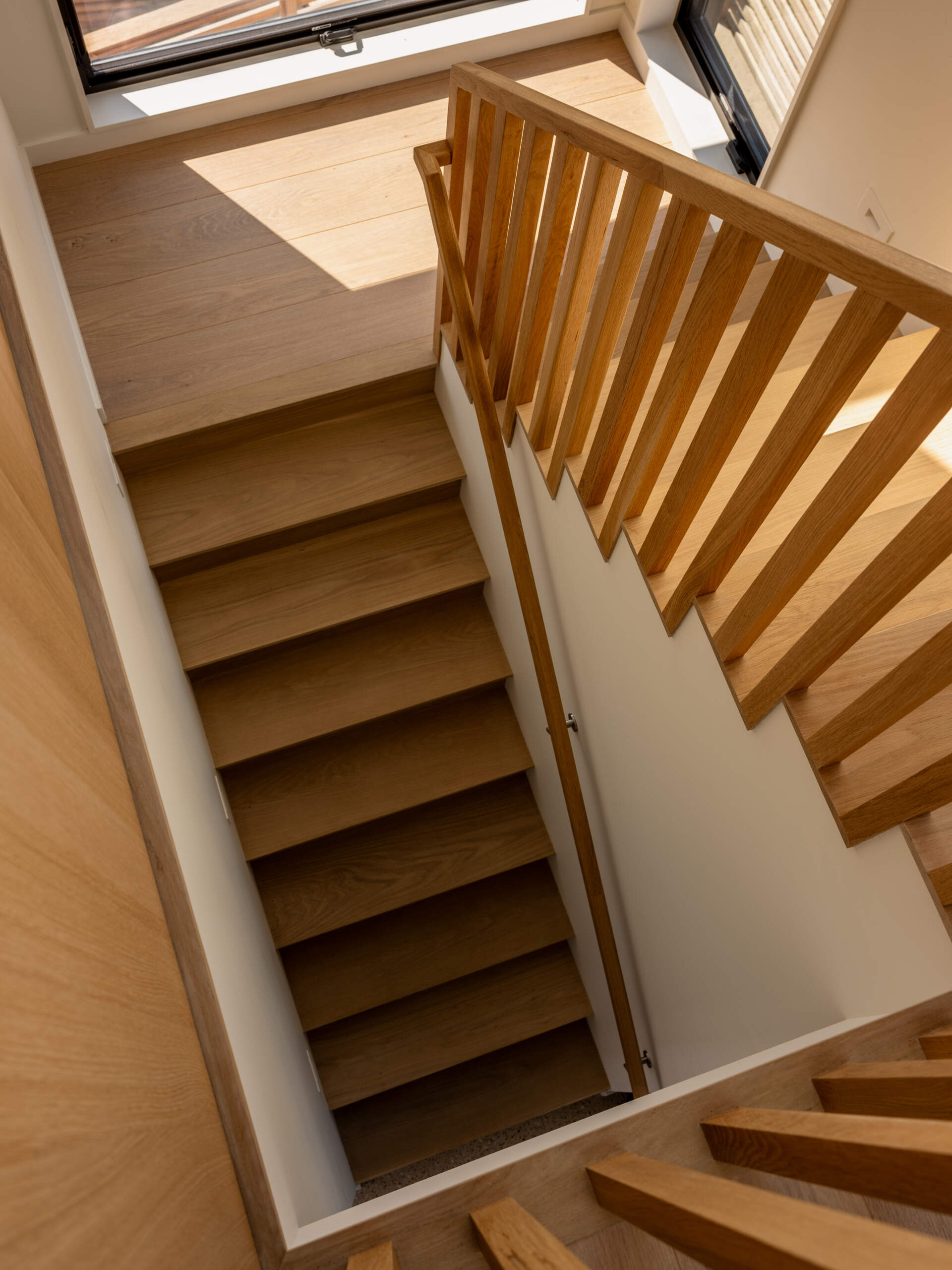 Timber steps, vertical wood balusters, and white walls create a simple, warm staircase in Surf Road House. Large black-framed windows illuminate the landing of this modern Fire Island home.