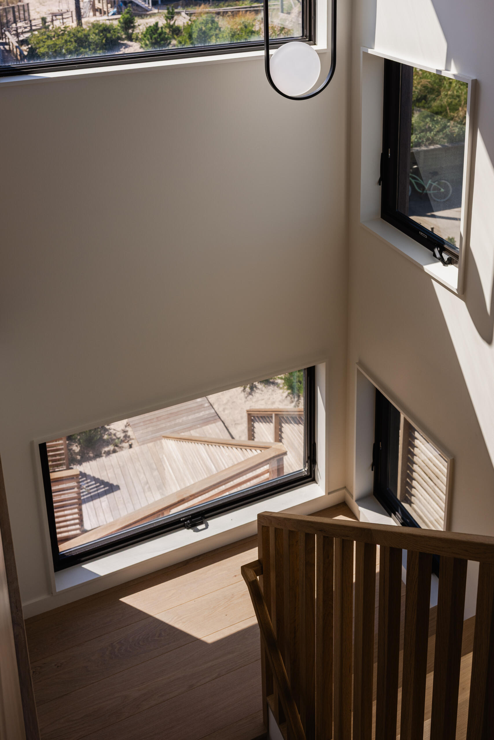Timber steps, vertical wood balusters, and white walls create a simple, warm staircase in Surf Road House. Large black-framed windows illuminate the landing of this modern Fire Island home.