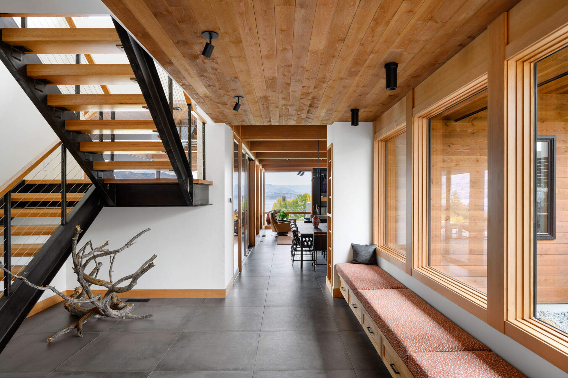A bright modern entryway combines warm timber, charcoal floor tiles, a built-in bench, and an open-riser staircase with black steel details.