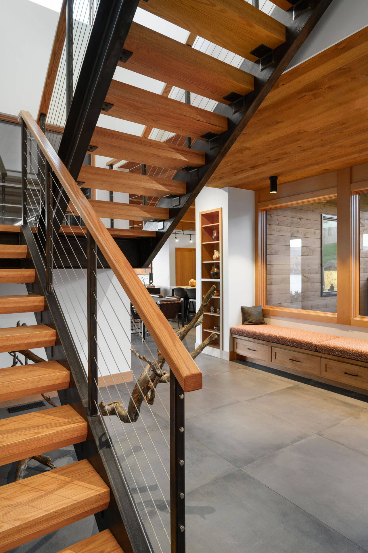 A dramatic staircase combines warm wood treads, black steel framing, cable railings, and built-in cabinetry, with a sculptural branch adding a natural detail.