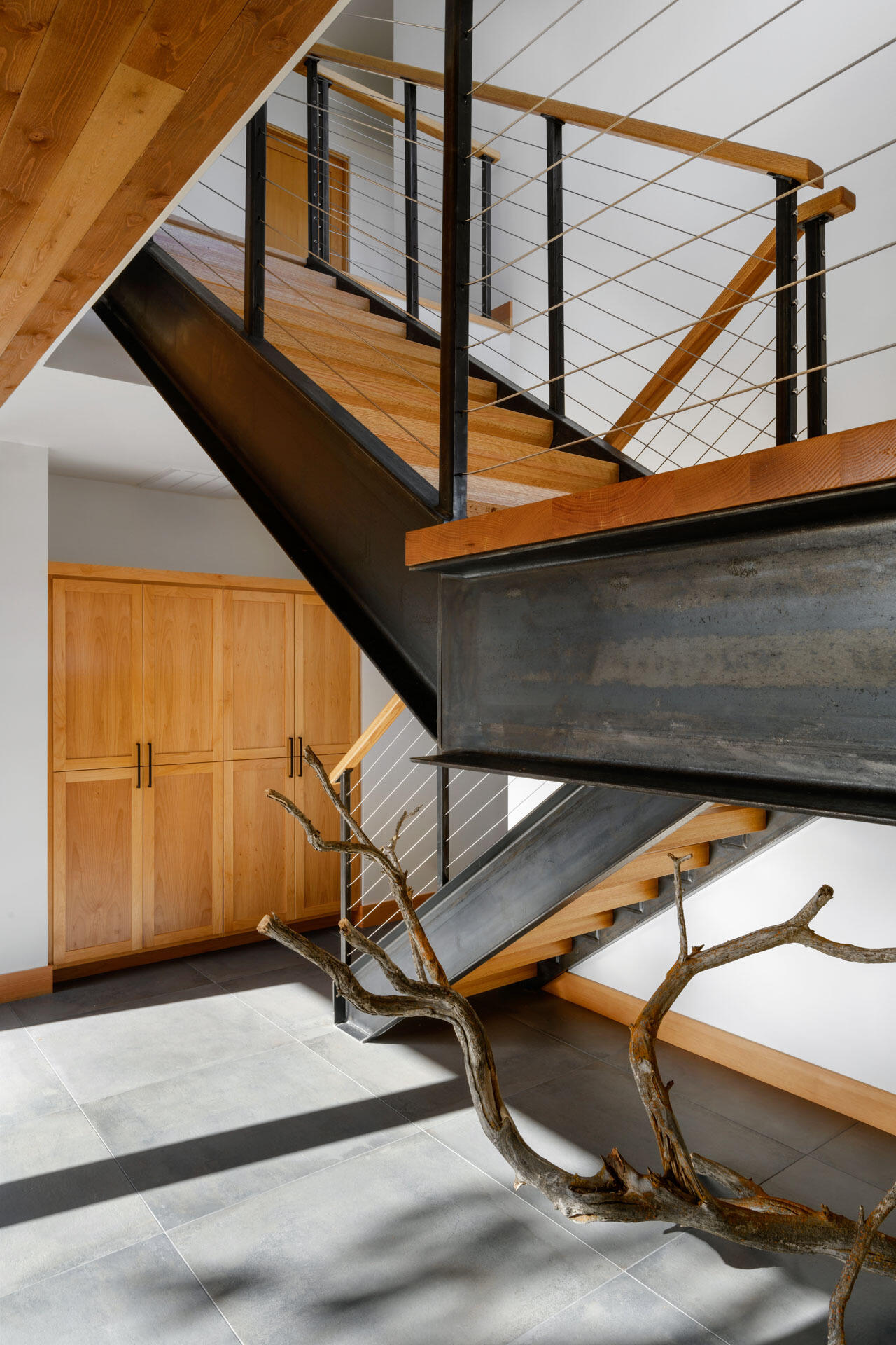 A dramatic staircase combines warm wood treads, black steel framing, cable railings, and built-in cabinetry, with a sculptural branch adding a natural detail.