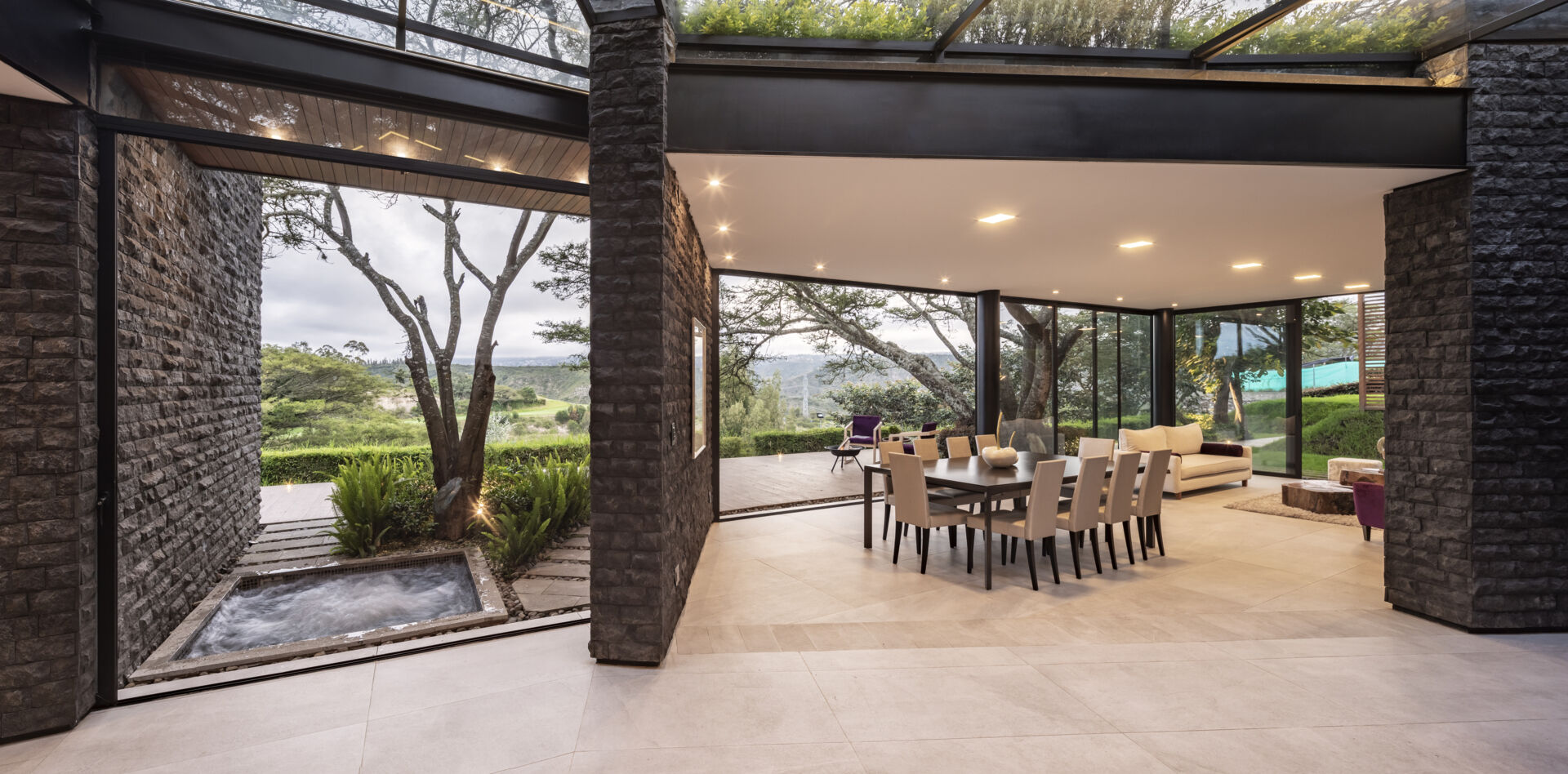 Explore the dining area of the Hidden Stone House, featuring a large dining table, light chairs, dark stone walls and views of the Ecuadorian landscape.