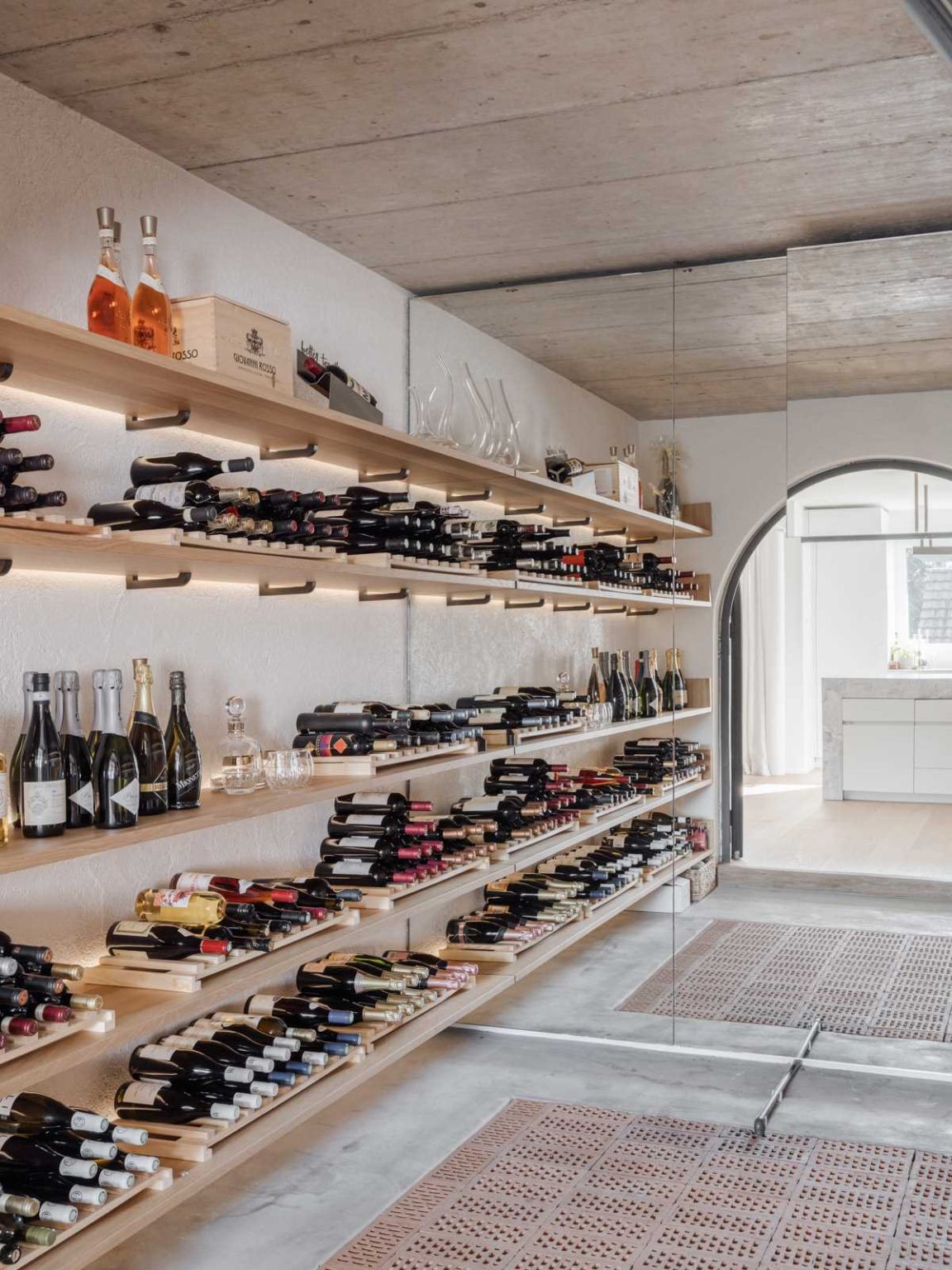 A beautiful Swiss wine cellar sits behind an arched black-framed glass doorway, with wood bottle storage, hidden lighting and a mirror that makes the compact space feel larger.