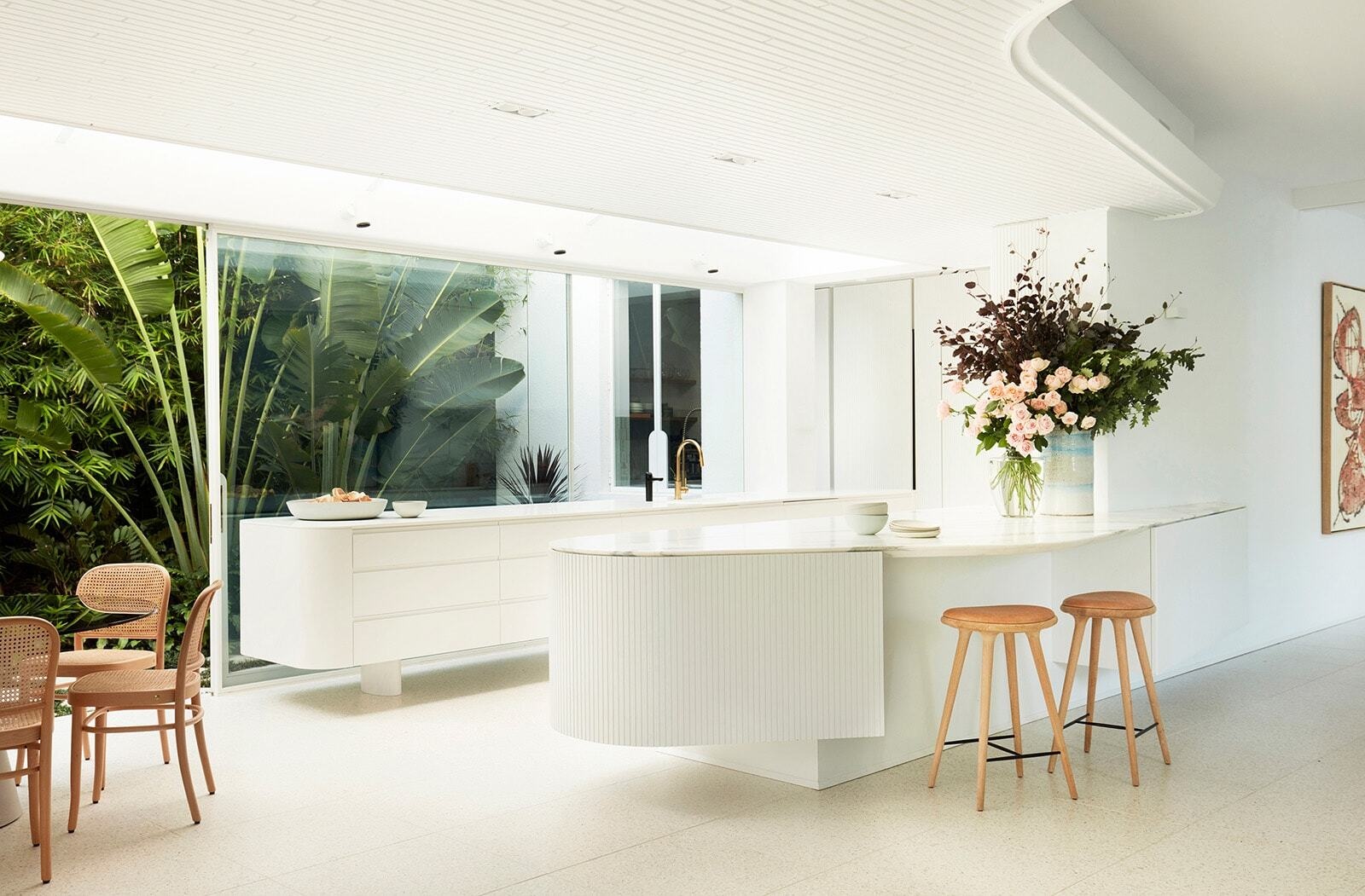 A light-filled open-plan kitchen, breakfast table and dining area sit beneath curved ceilings, with expansive glass opening the home toward its lush garden.
