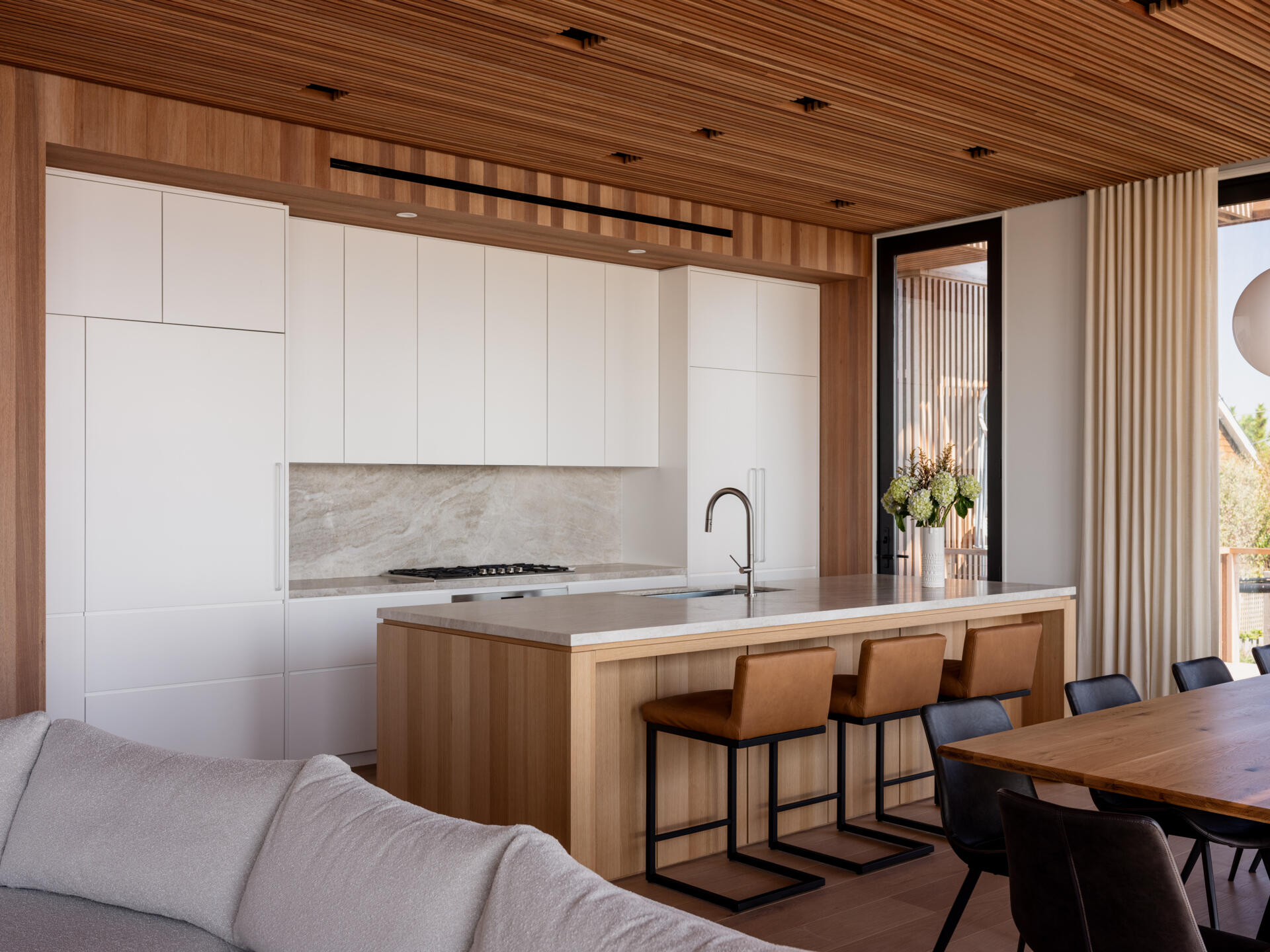 White cabinetry, a wood kitchen island, and tan counter stools meet a long dining table in this open-plan Fire Island home. Globe pendants and trellis-filtered sunlight add character, with sliding doors leading to outdoor dining.