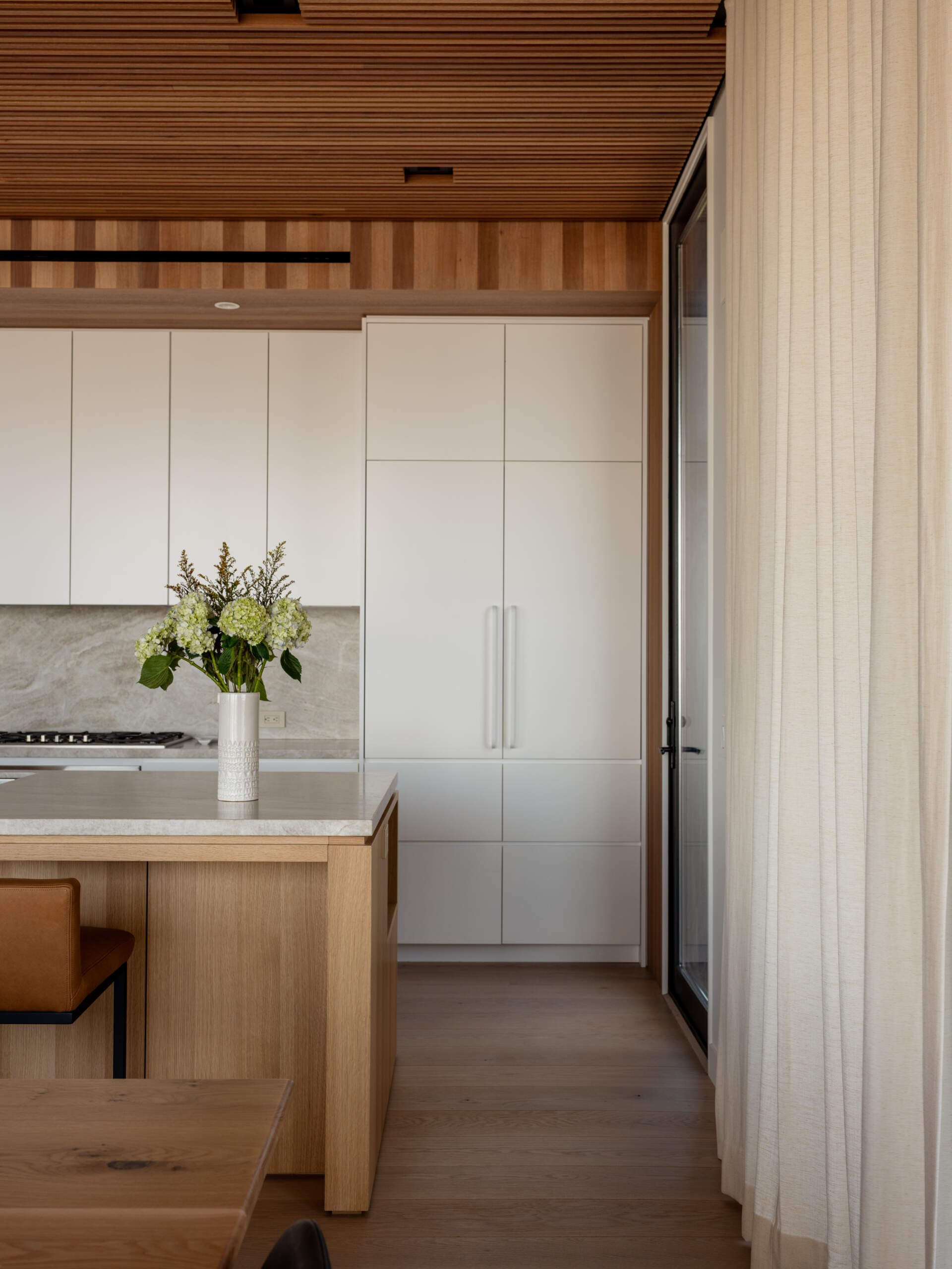 White cabinetry, a wood kitchen island, and tan counter stools meet a long dining table in this open-plan Fire Island home. Globe pendants and trellis-filtered sunlight add character, with sliding doors leading to outdoor dining.