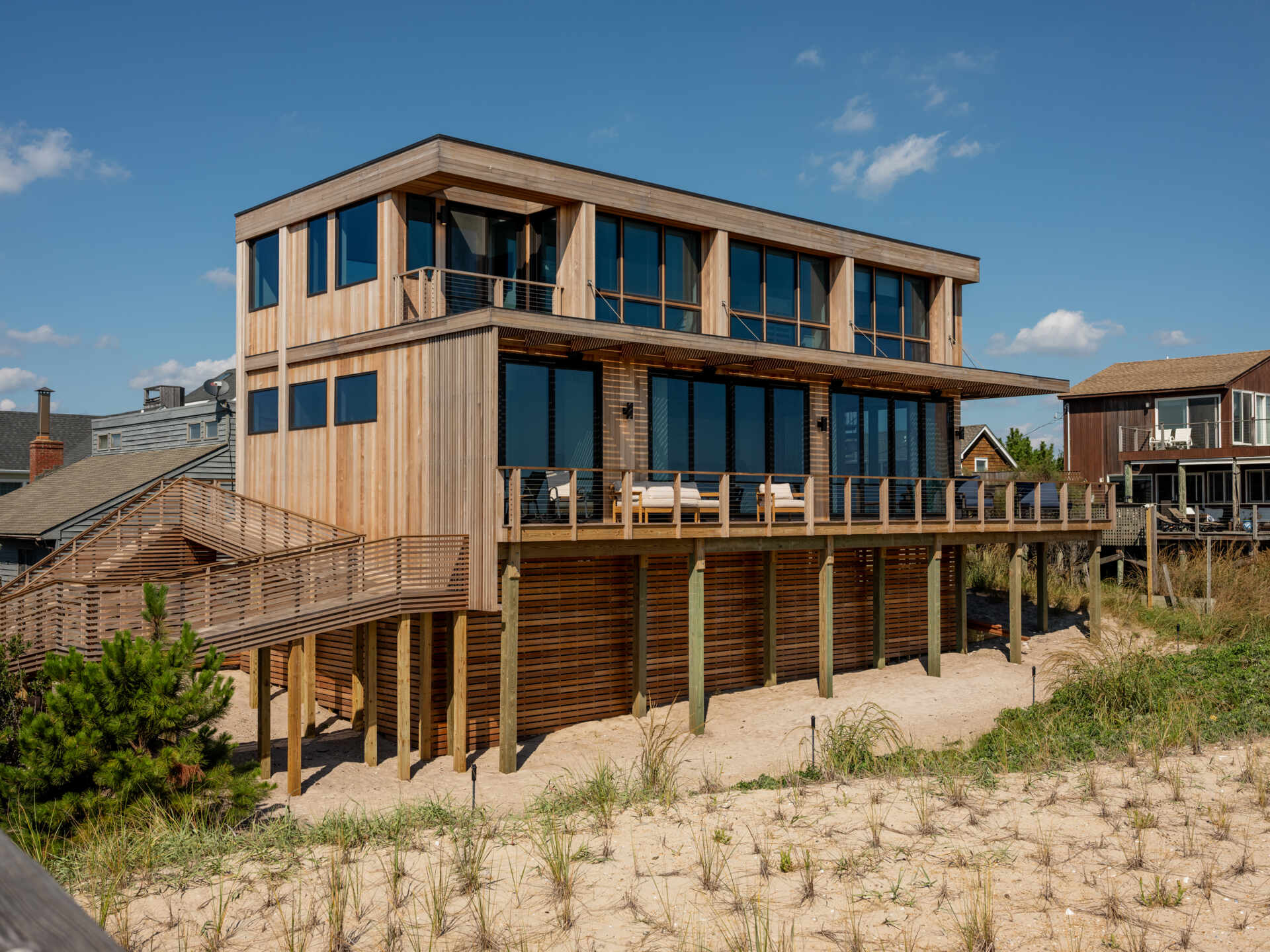 This ocean-facing balcony at Surf Road House pairs timber lounge chairs with a slatted wood trellis and cable railing. Opening from the living room, it overlooks the dunes, boardwalk, and beach on Fire Island.