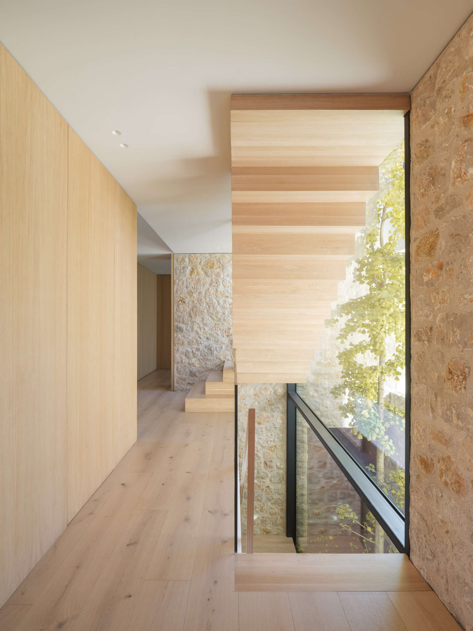 A sculptural wooden staircase with a zigzag profile, glass balustrade, and timber handrail rises beside a planted vertical patio. Explore the light-filled stairwell at House 2194 by Nordest Architecture SLP in Olot, Spain.