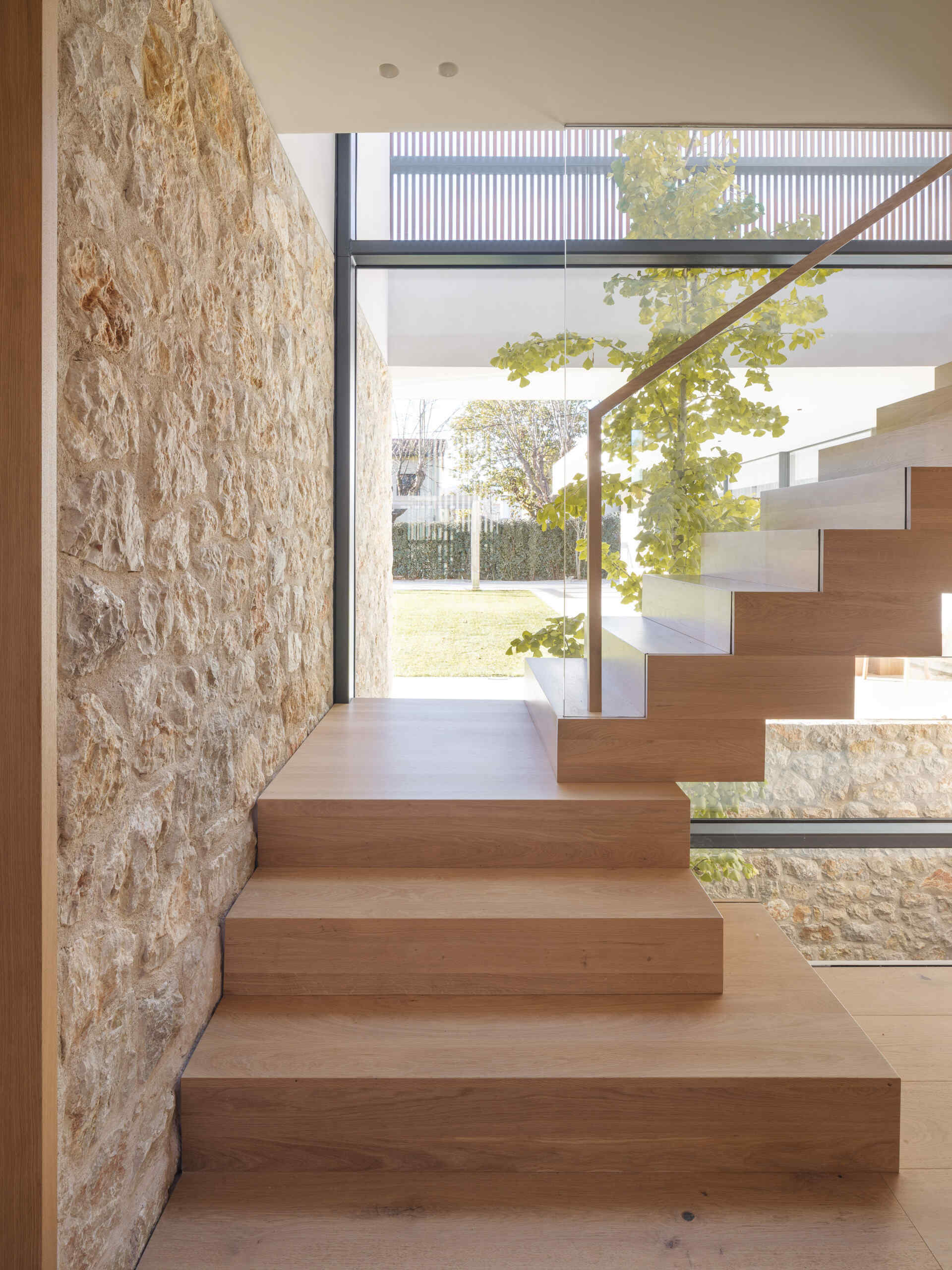 A sculptural wooden staircase with a zigzag profile, glass balustrade, and timber handrail rises beside a planted vertical patio. Explore the light-filled stairwell at House 2194 by Nordest Architecture SLP in Olot, Spain.