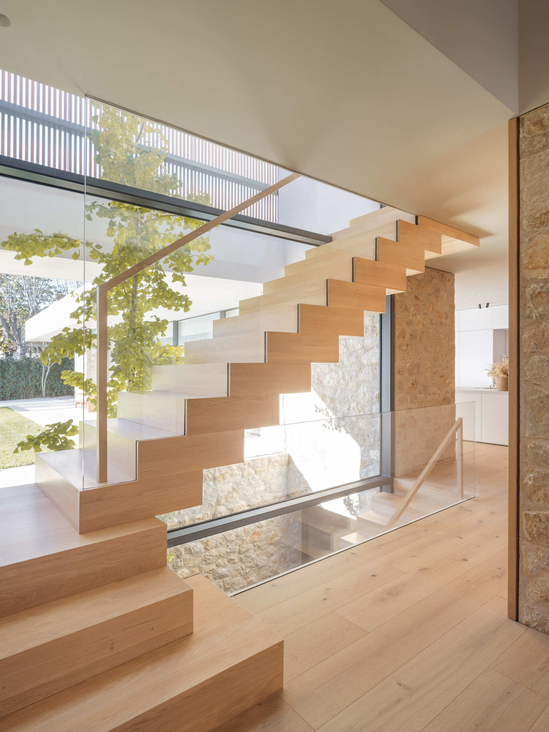 A sculptural wooden staircase with a zigzag profile, glass balustrade, and timber handrail rises beside a planted vertical patio. Explore the light-filled stairwell at House 2194 by Nordest Architecture SLP in Olot, Spain.