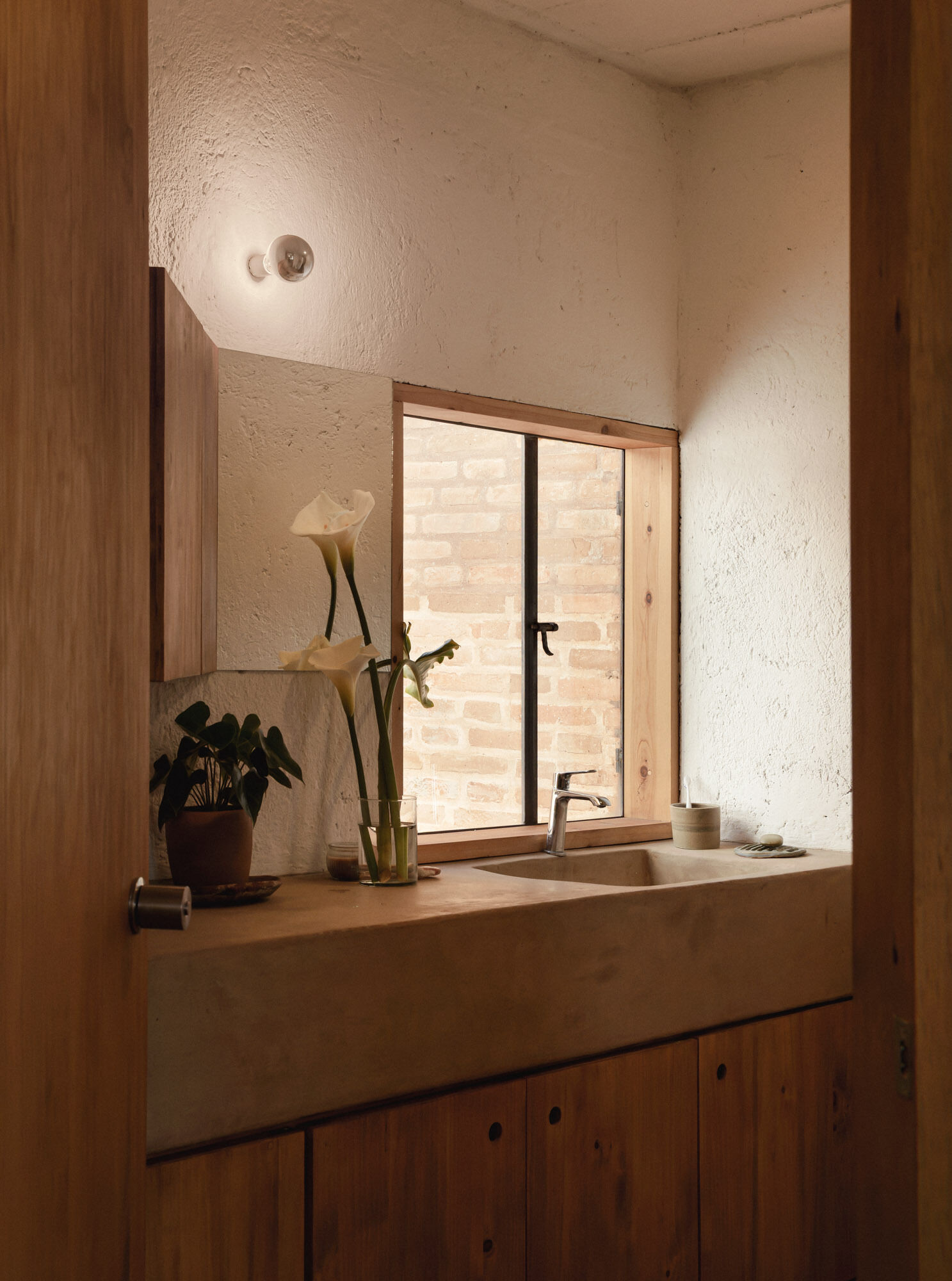 Casa Amona’s bathroom pairs a built-in basin and cypress storage with a window overlooking exposed brick and a small planted space.